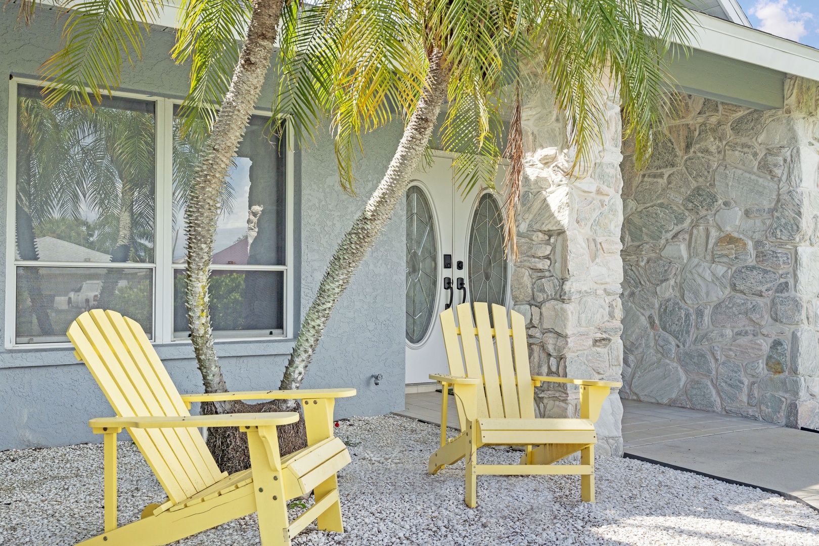 Mid-century modern charm meets desert oasis living. Two cheerful yellow chairs invite you to relax beneath swaying palms, framing the entrance to your stylish escape with its vintage stone accents and retro elegance.