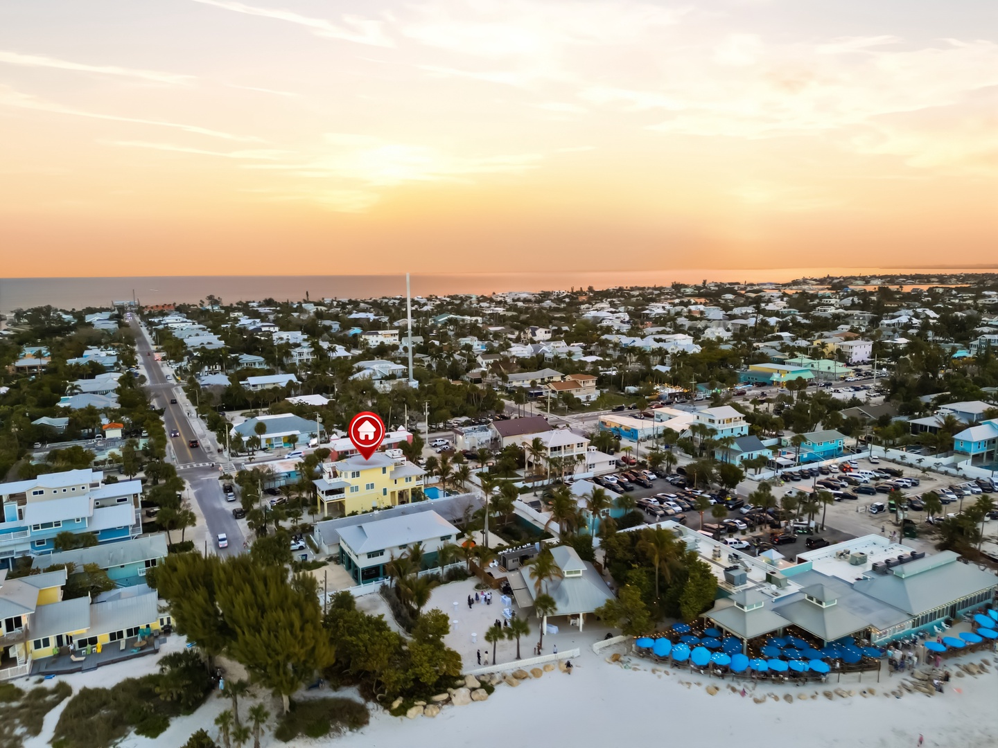 Aerial view of a vibrant coastal community at sunset, with colorful beach houses nestled among palm trees near pristine white sand beaches.