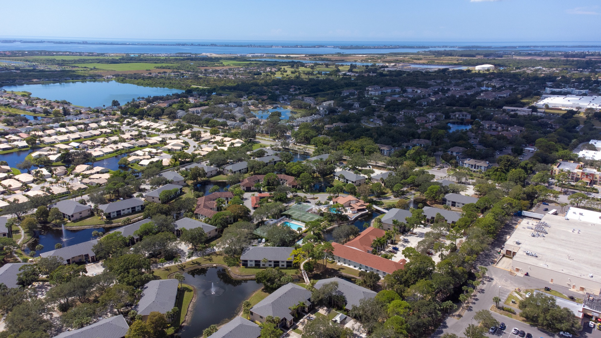Aerial view of residential neighborhood with lakefront homes, swimming pools, and tree-lined streets extending toward coastal waters.