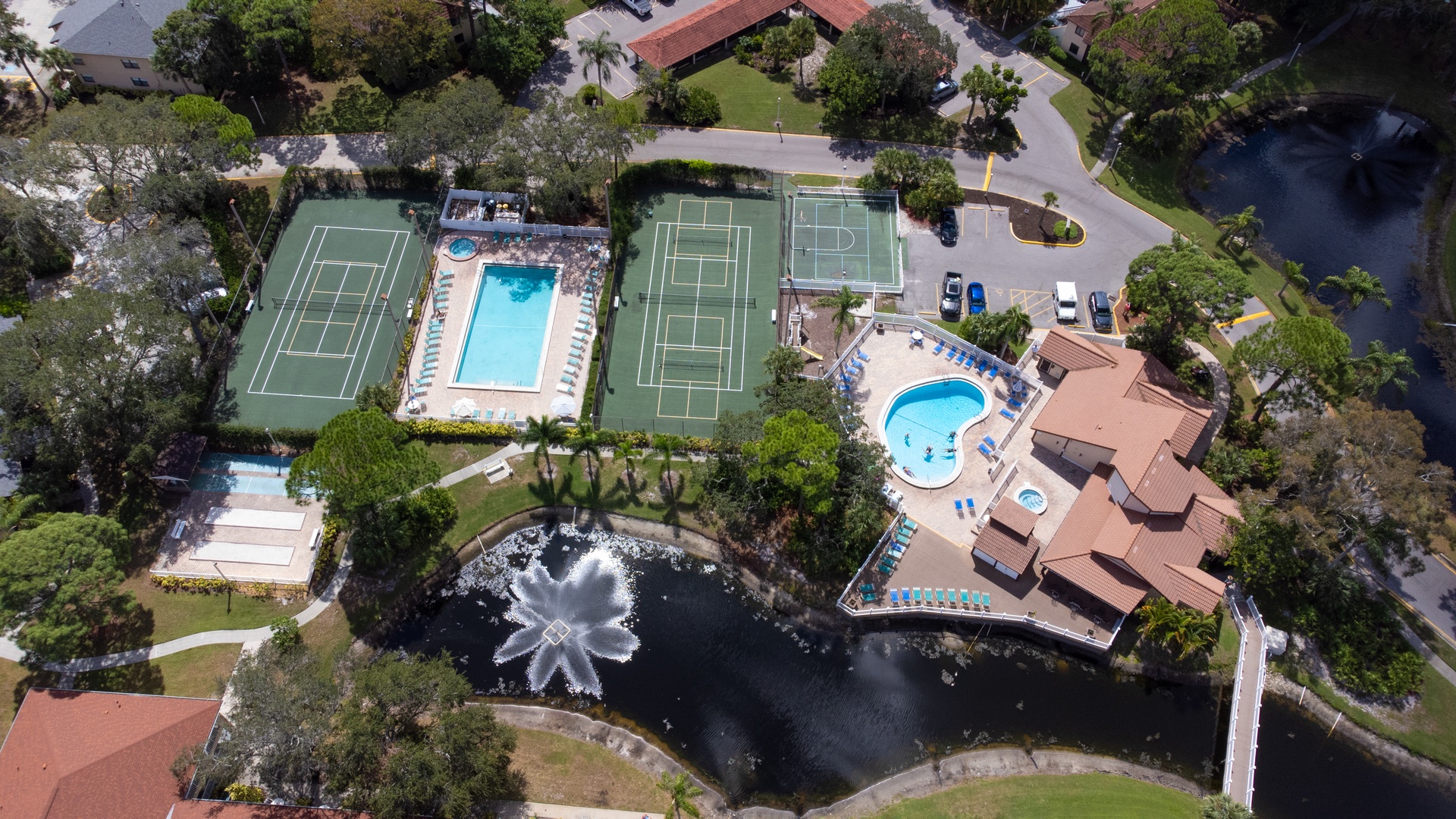 Aerial view of resort-style amenities featuring tennis courts, swimming pools, and recreational facilities surrounded by lush landscaping and water features.