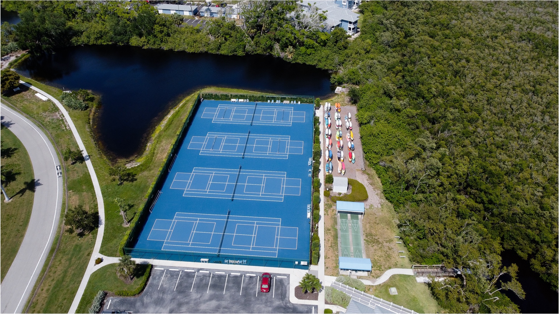 Aerial view of tennis courts adjacent to a tranquil lake, surrounded by lush natural landscape and nearby residential areas.