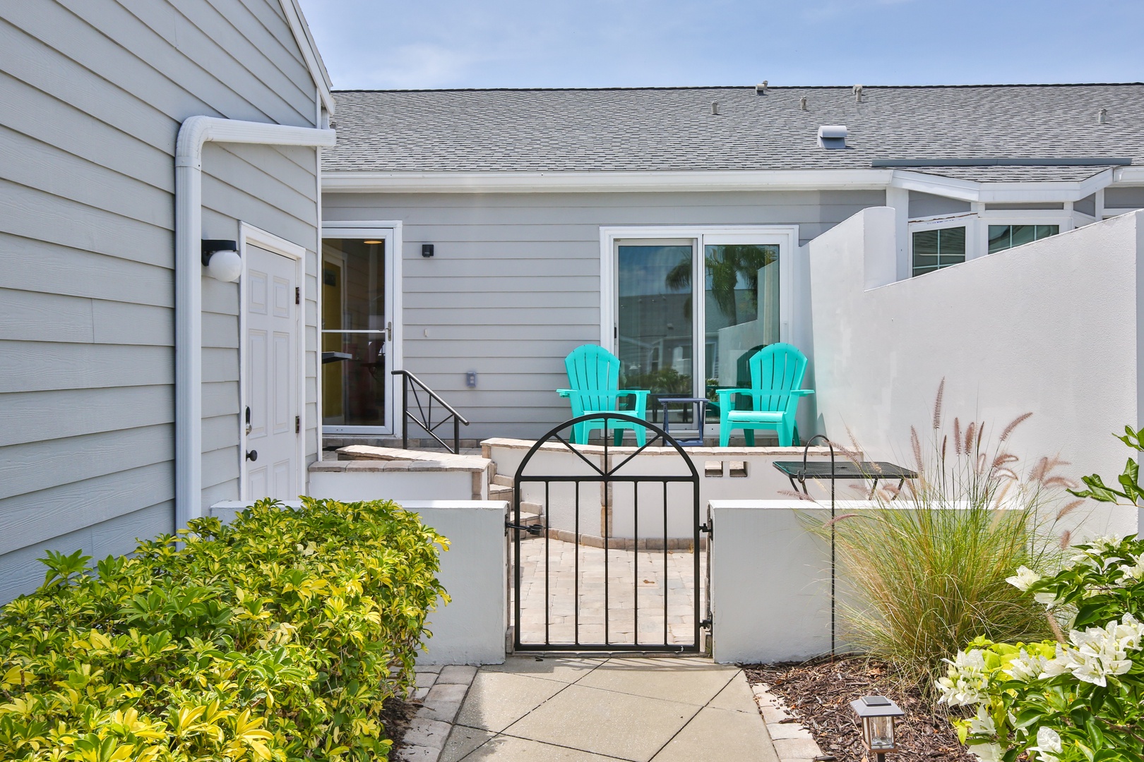 Charming property entrance with a welcoming gate opening to a private patio featuring turquoise Adirondack chairs, perfect for relaxing mornings or sunset evenings outdoors.