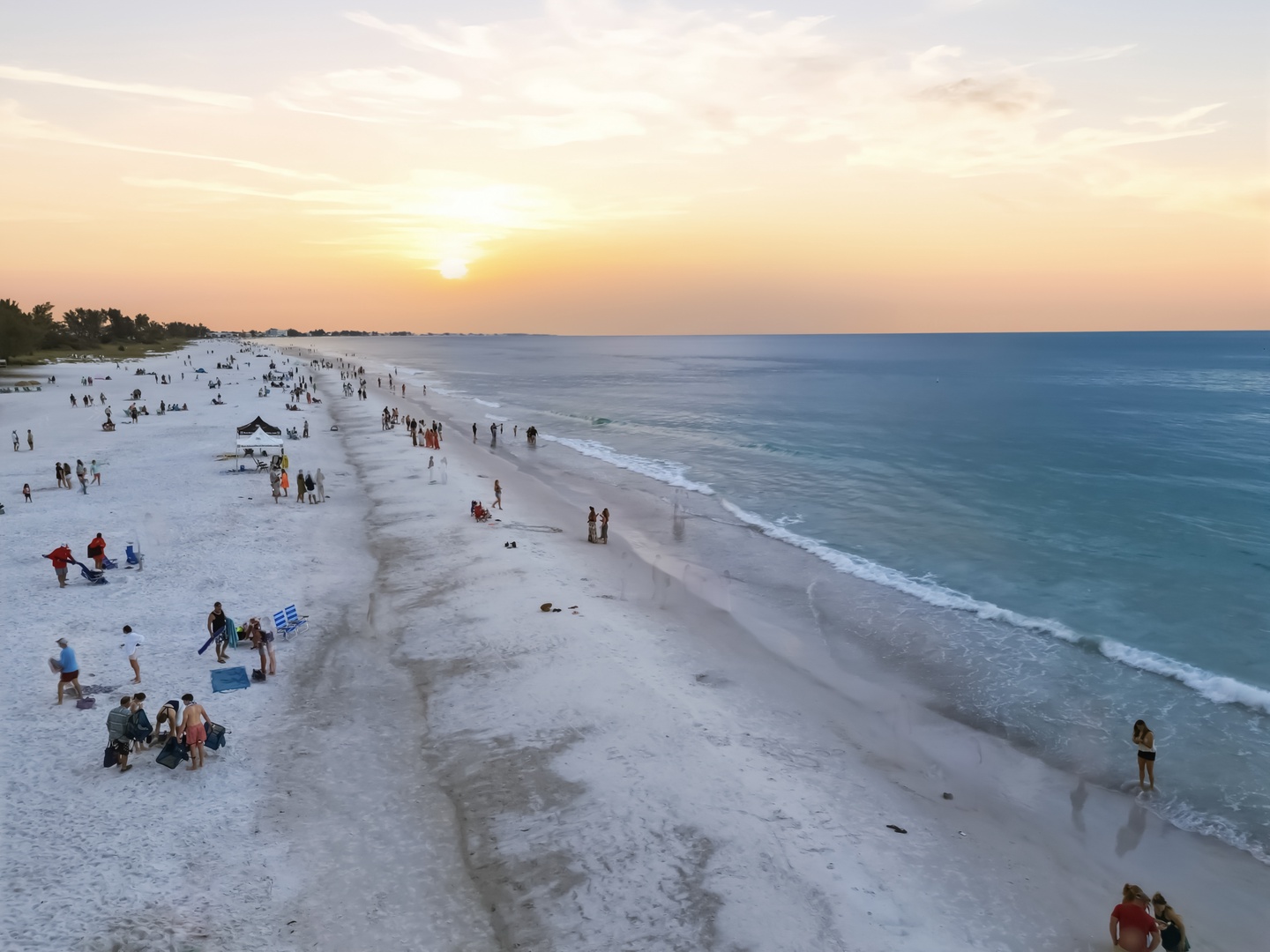 Pristine white sand beach stretches along crystal-clear waters, with visitors enjoying the stunning sunset over the Gulf Coast.