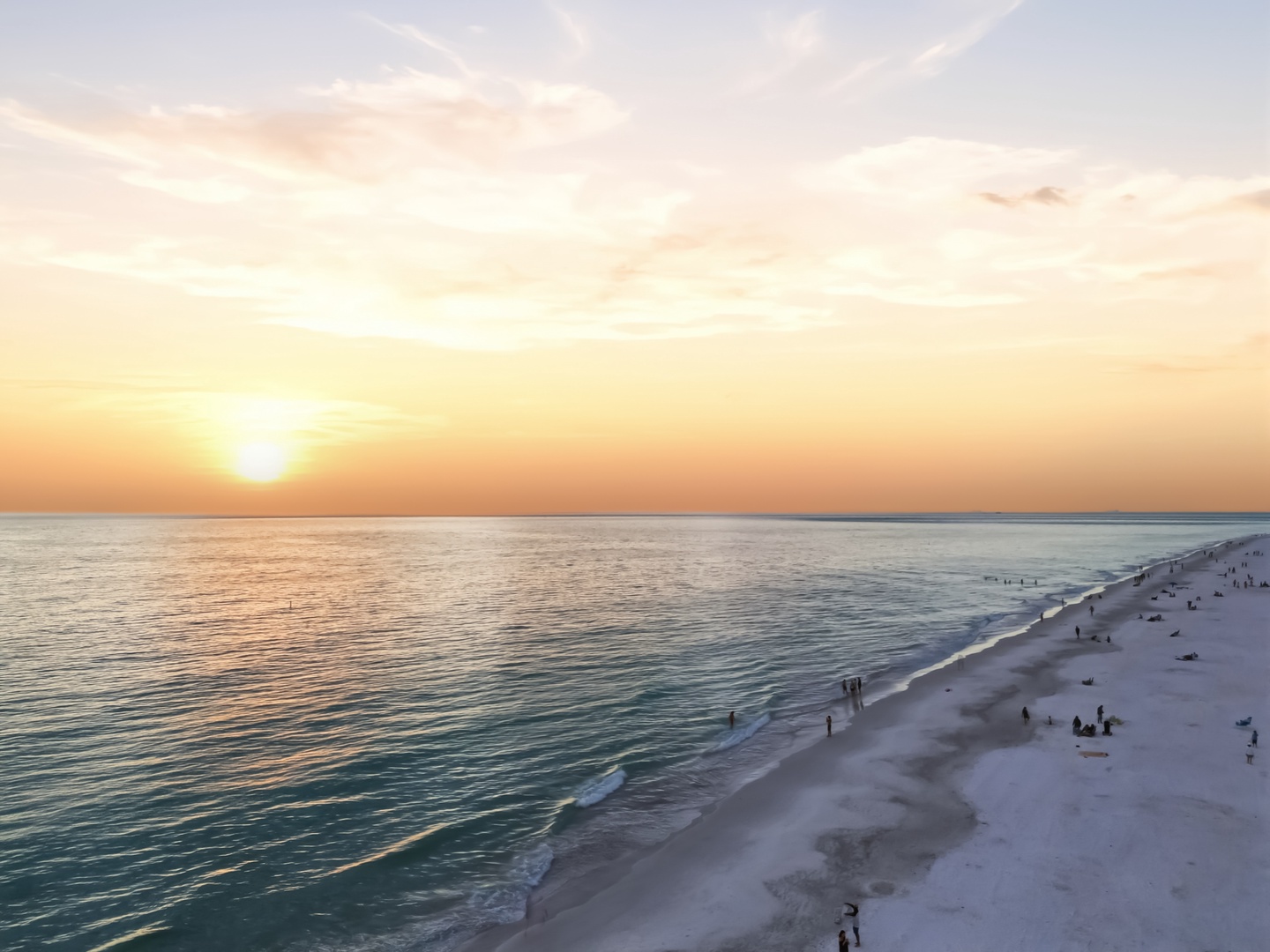 Golden sunset paints the sky over pristine white sand beach with gentle waves and scattered beachgoers enjoying the coastal paradise.