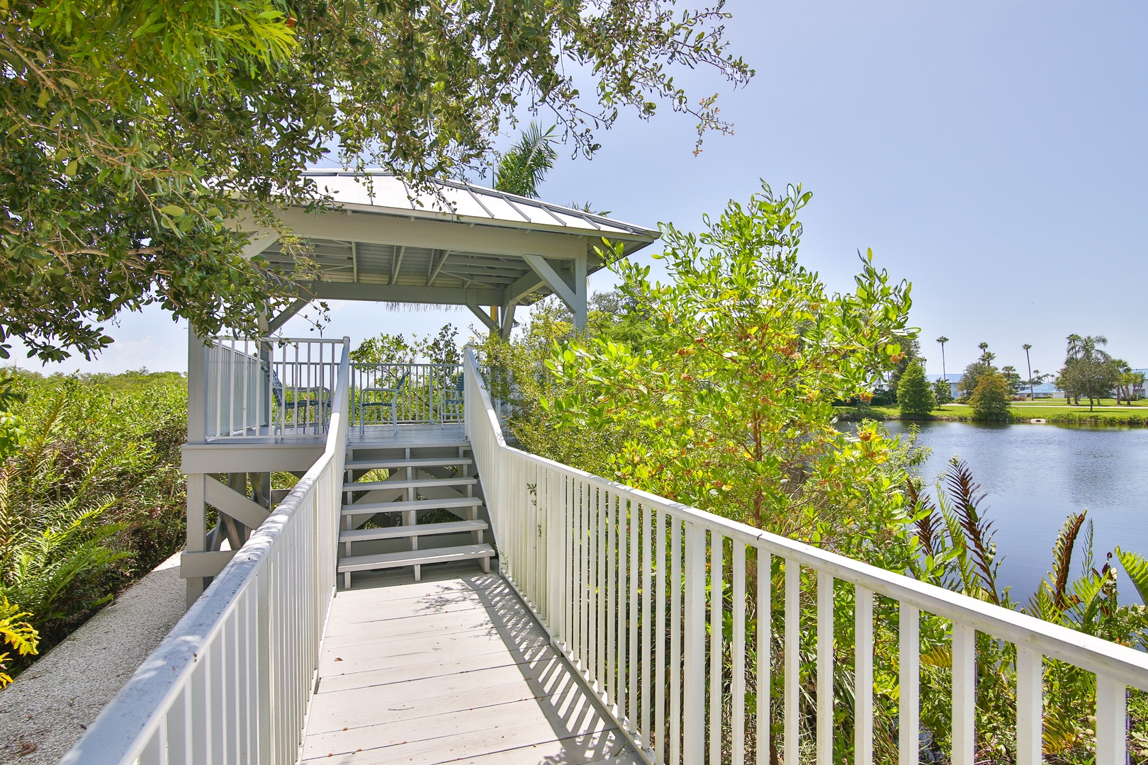 A covered gazebo overlooks tranquil water, surrounded by lush tropical landscaping and accessed via a charming wooden walkway—part of the property's scenic outdoor surroundings.