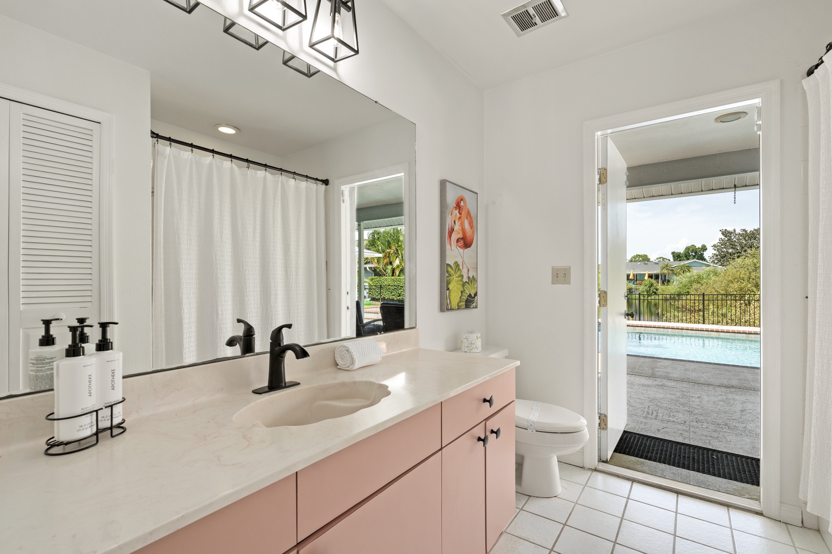 Step into your private oasis where soft pink cabinetry and chic black fixtures create a spa-like retreat. Natural light pours in as French doors frame views of your sparkling pool and lush surroundings.