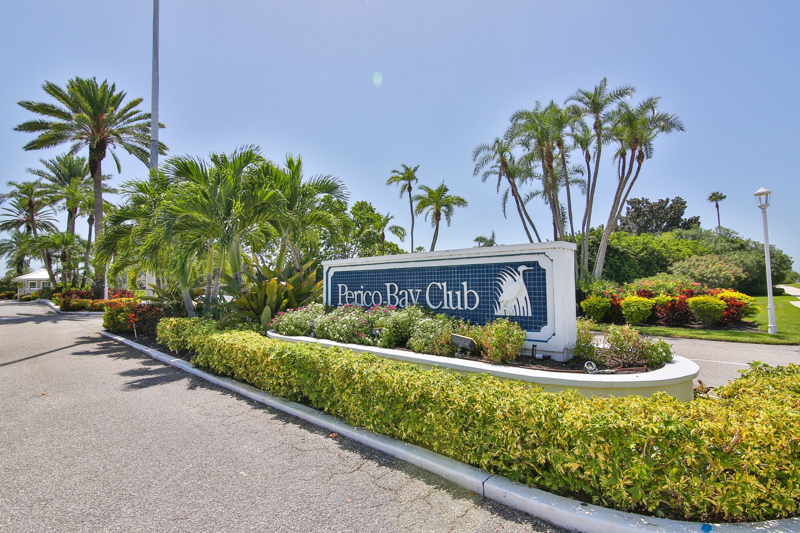 The entrance to Perico Bay Club, surrounded by lush tropical landscaping and vibrant flowering plants under clear blue skies.