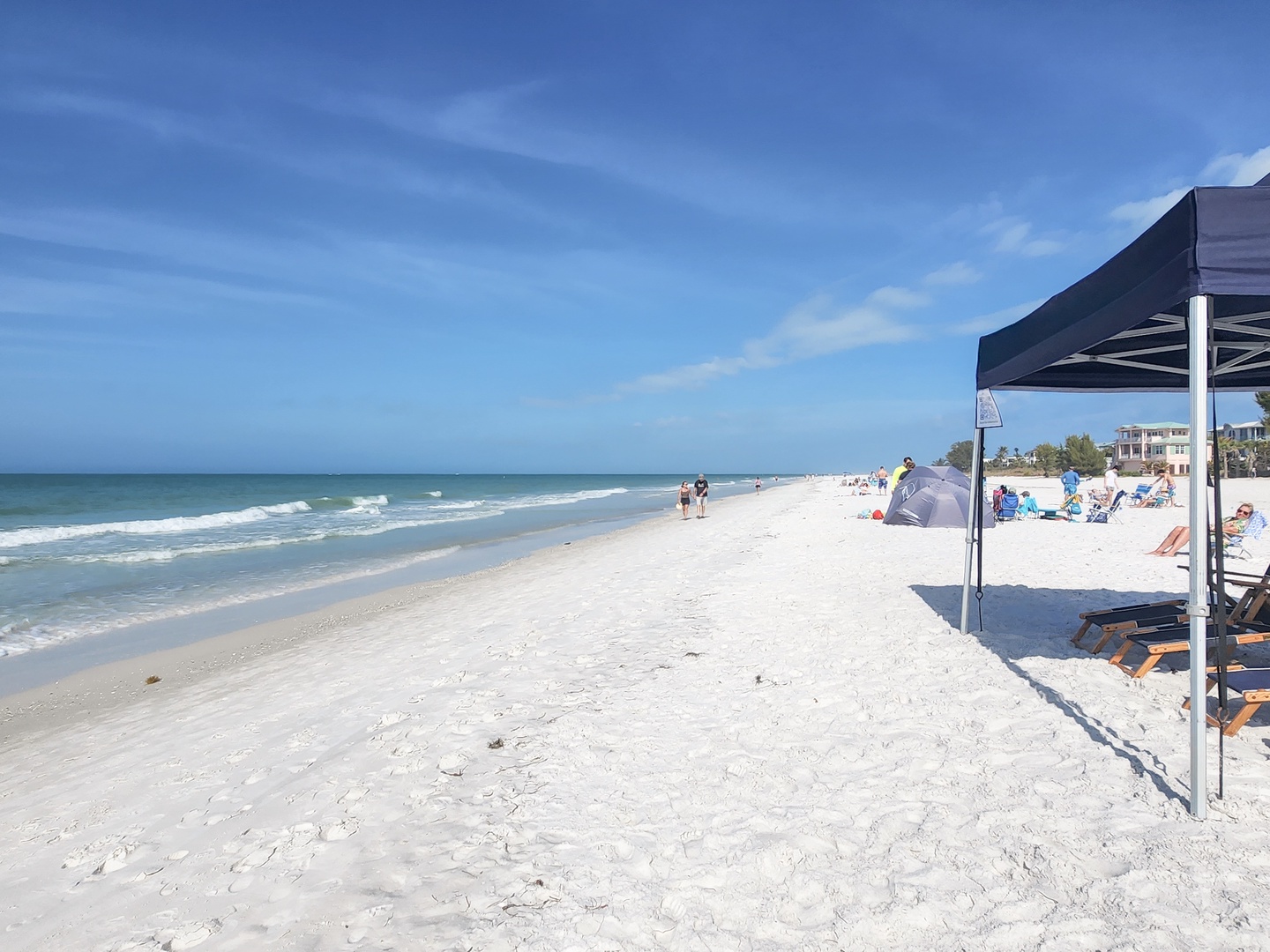 Pristine white sand beach stretches along turquoise waters, with comfortable shade structures and nearby waterfront properties visible in the distance.