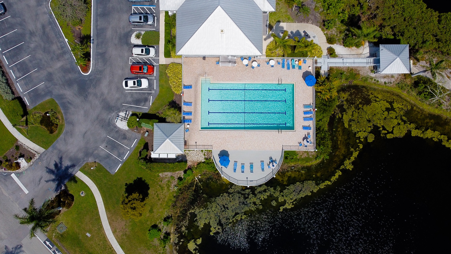 Aerial view showcasing the community swimming pool, poolside deck with lounge chairs, and natural lakeside setting framed by lush greenery and convenient parking.