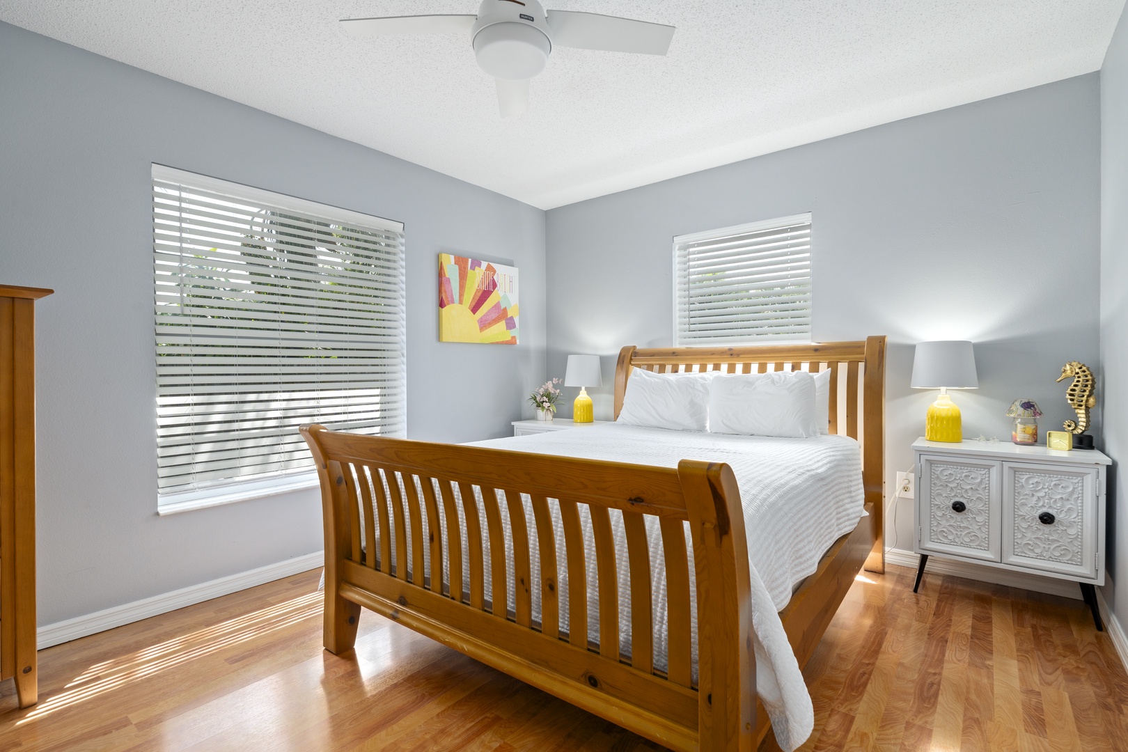 Sink into this cozy bedroom where soothing gray-blue walls meet warm wood tones. Your restful nights begin with crisp linens and gentle sunlight filtered through plantation shutters, all cooled by a quiet ceiling fan above.