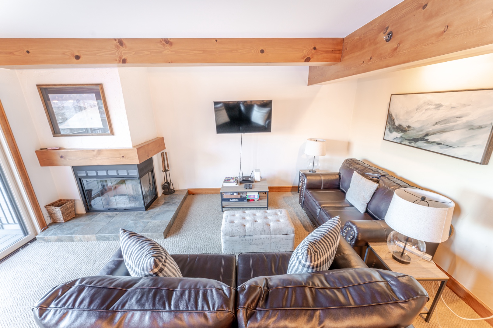 A cozy living room features a lit fireplace, two brown sofas, a large wall-mounted TV, and artworks on the walls. Exposed wooden beams add a rustic touch.