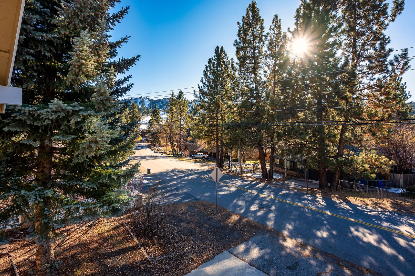Sunny view of Bear Mountain Ski Resort featuring tree-lined residential street with snowy patches set against the ski slopes..