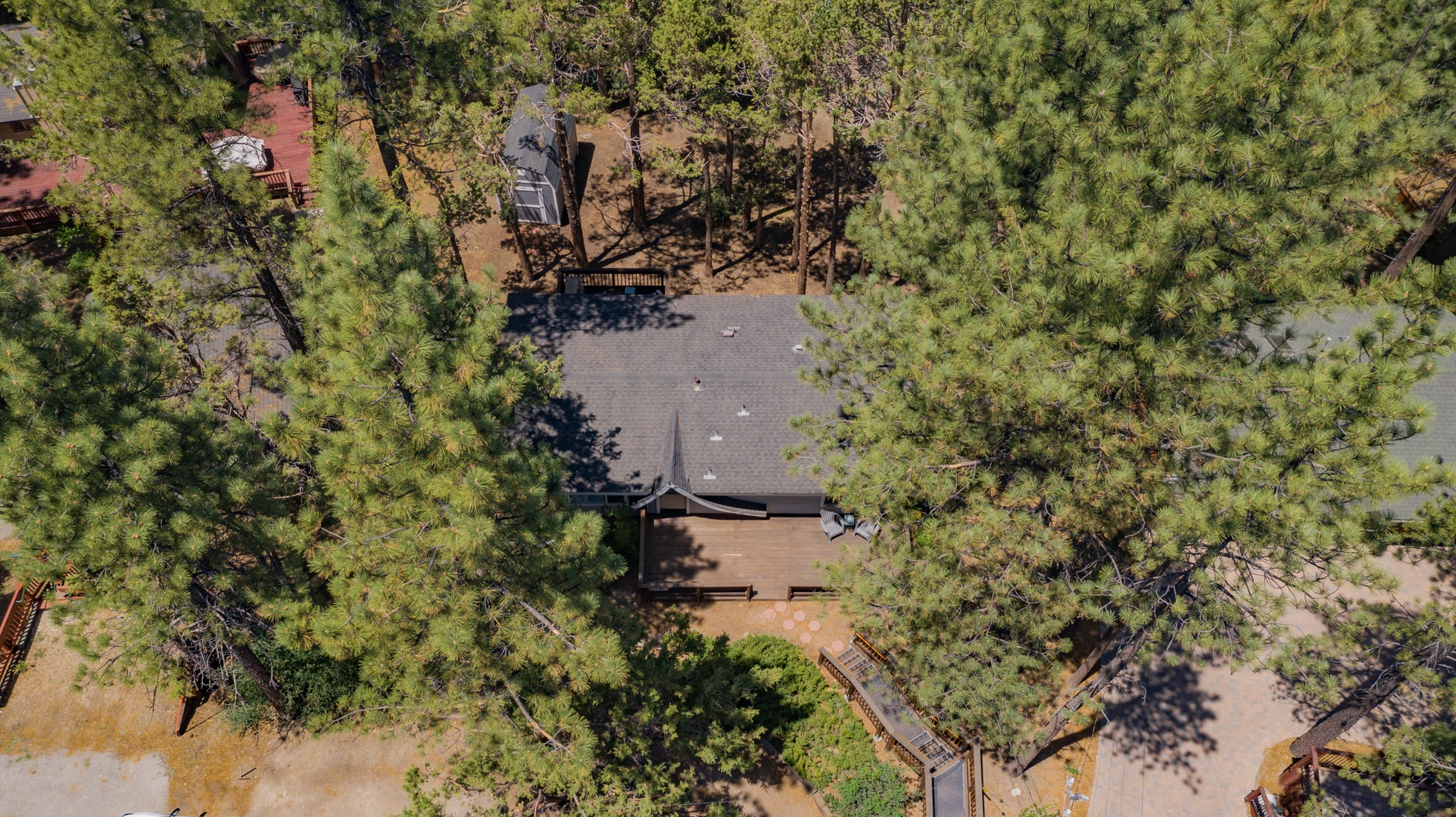 Aerial view of a charming vacation cabin nestled among towering pines. The property features a classic gray roof and private deck, surrounded by lush forest and neighboring homes.
