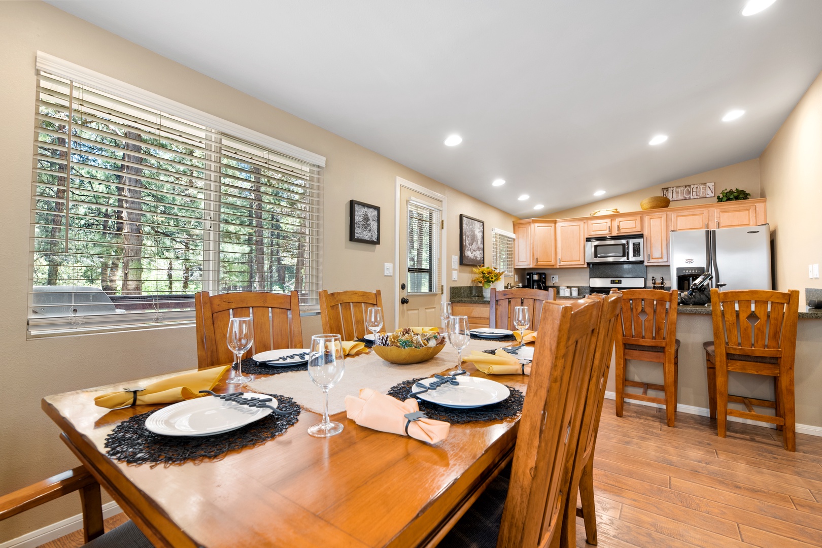 Gather around this inviting dining table, beautifully set and ready for your family meals. The open layout flows seamlessly into a fully equipped kitchen, with natural light streaming through large windows overlooking lush greenery.