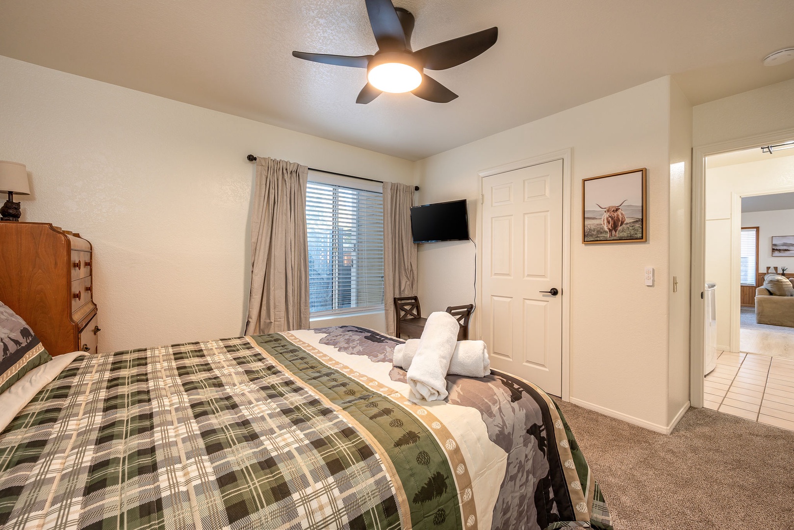Relax in this comfortable bedroom featuring a cozy plaid bedspread, ceiling fan for optimal comfort, and rustic highland cow artwork that adds character to your stay.