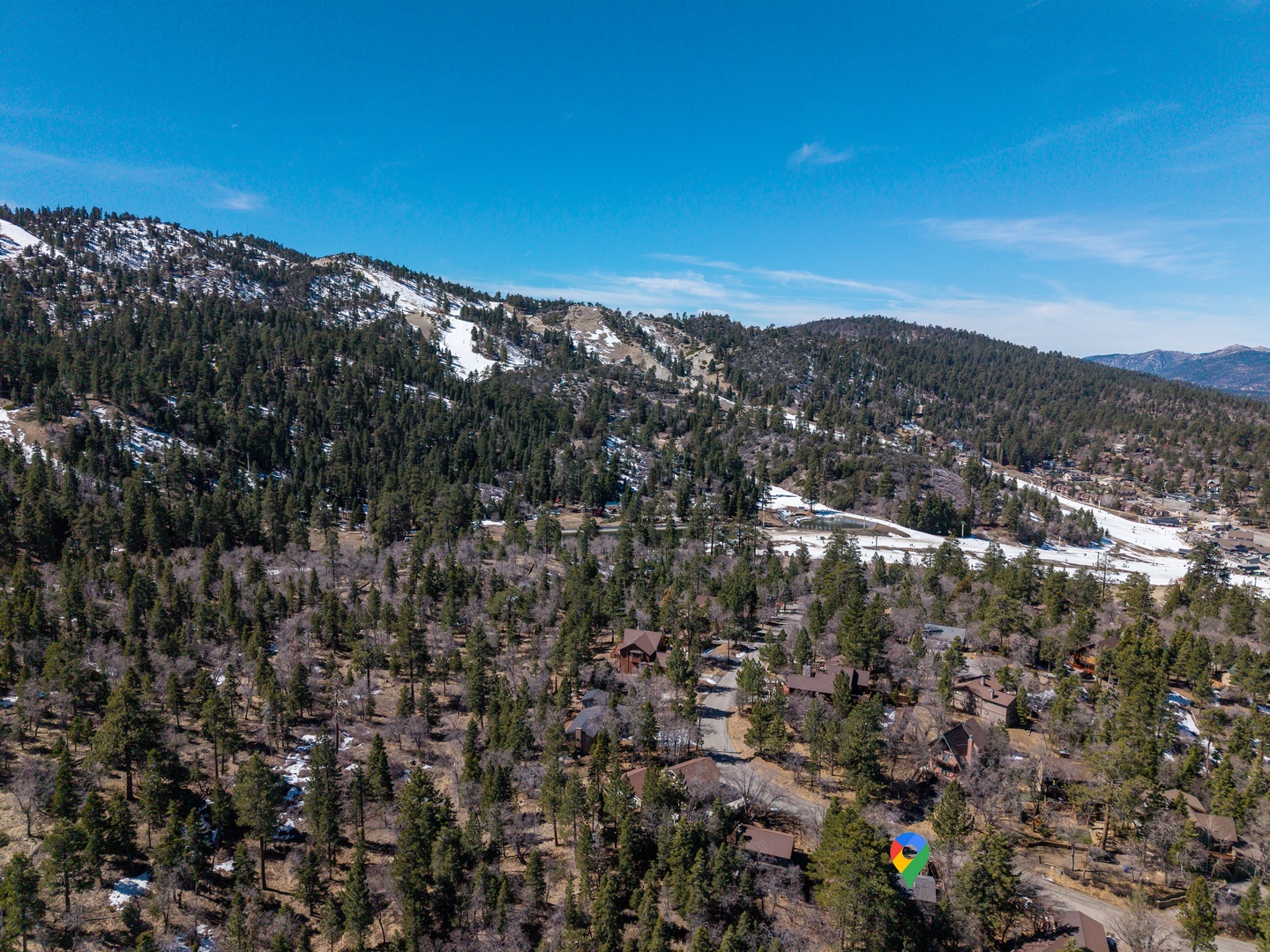 Aerial view of mountain community nestled among snow-dusted peaks and dense evergreen forests under brilliant blue skies.
