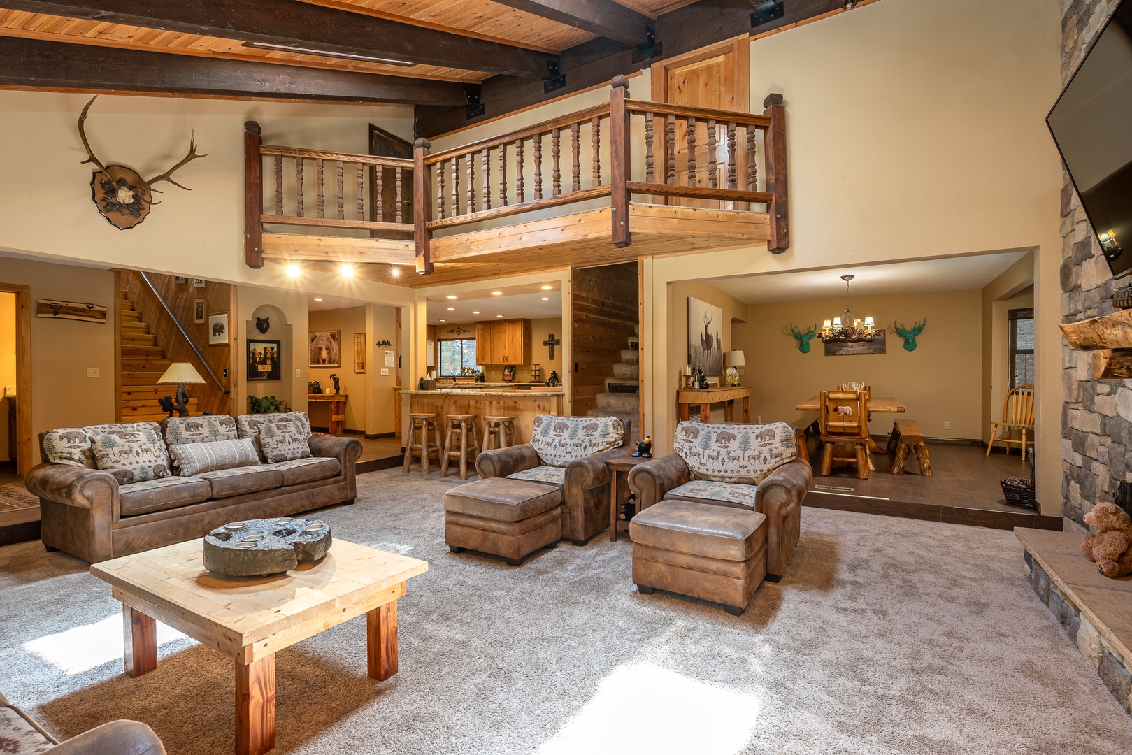 Sink into plush leather seating beneath soaring wood-beam ceilings in your spacious mountain retreat, where rustic charm meets modern comfort with a lofted balcony and open kitchen nearby.