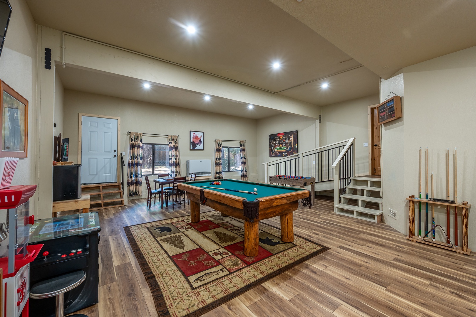 A spacious game room featuring a full-size pool table, arcade games, and dining area—perfect for friendly competition and entertainment. Natural light pours through large windows, highlighting polished hardwood floors and comfortable spaces to relax betwe