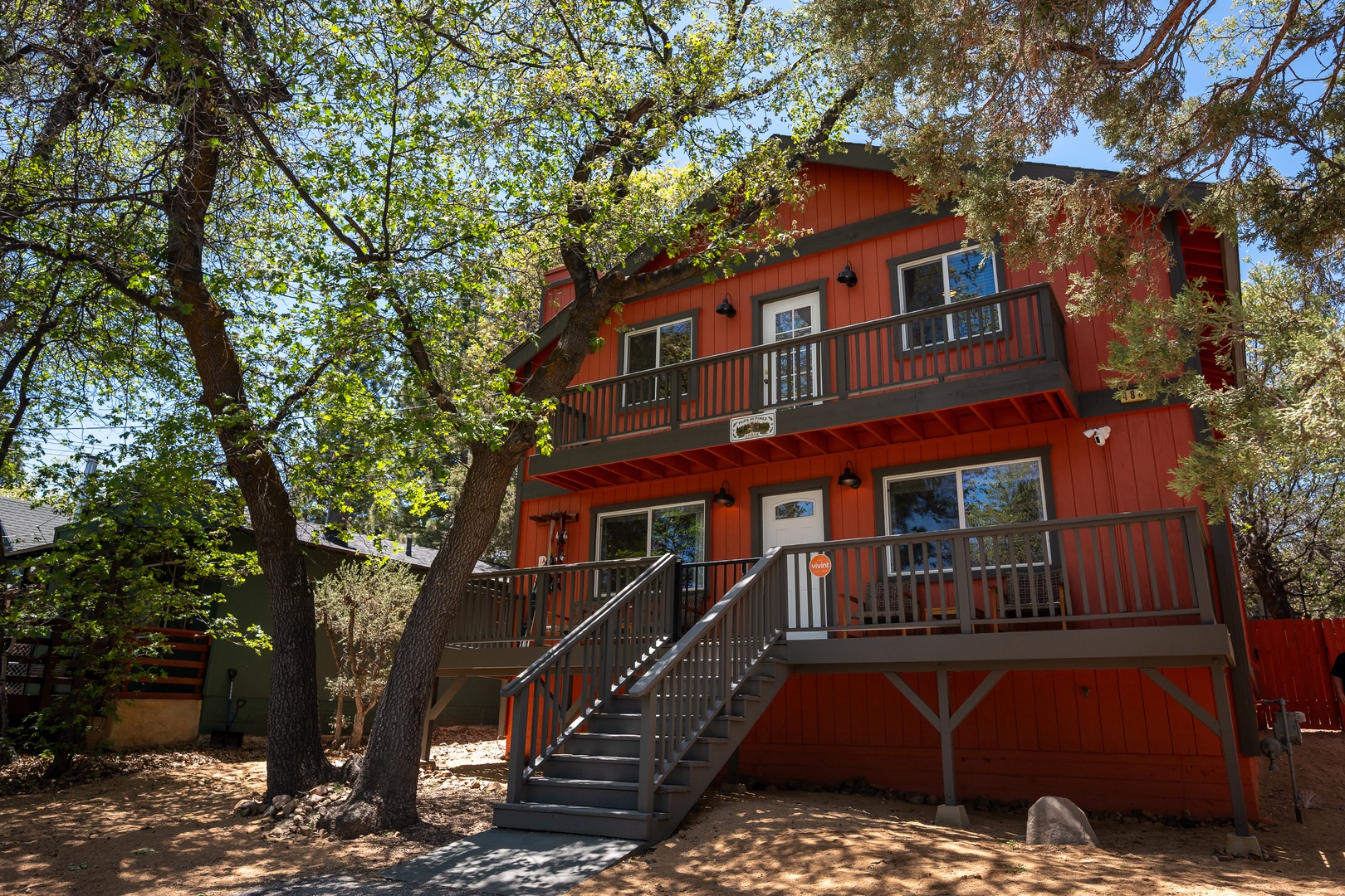 Charming red cabin-style property nestled among mature trees, featuring multi-level balconies and a welcoming stairway entrance. A peaceful retreat blending rustic character with natural surroundings in a wooded setting.
