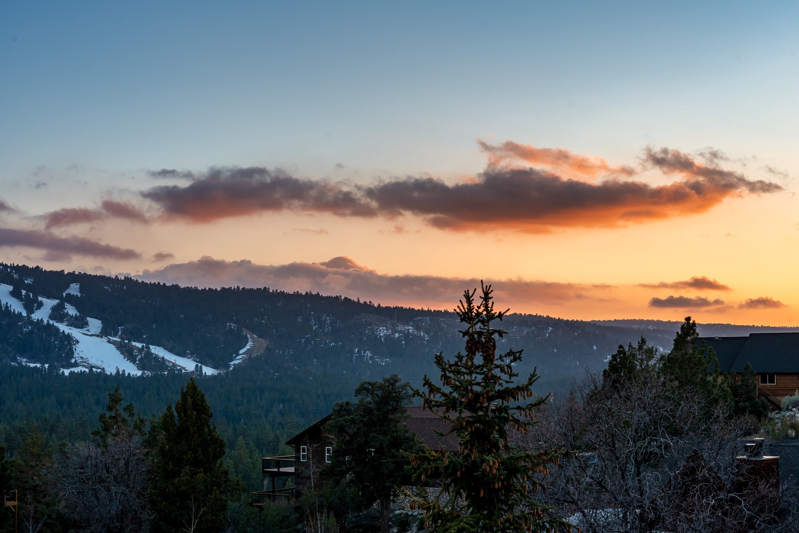 Stunning mountain sunset paints the sky in warm hues above snow-dusted peaks and forested slopes in this picturesque alpine setting.