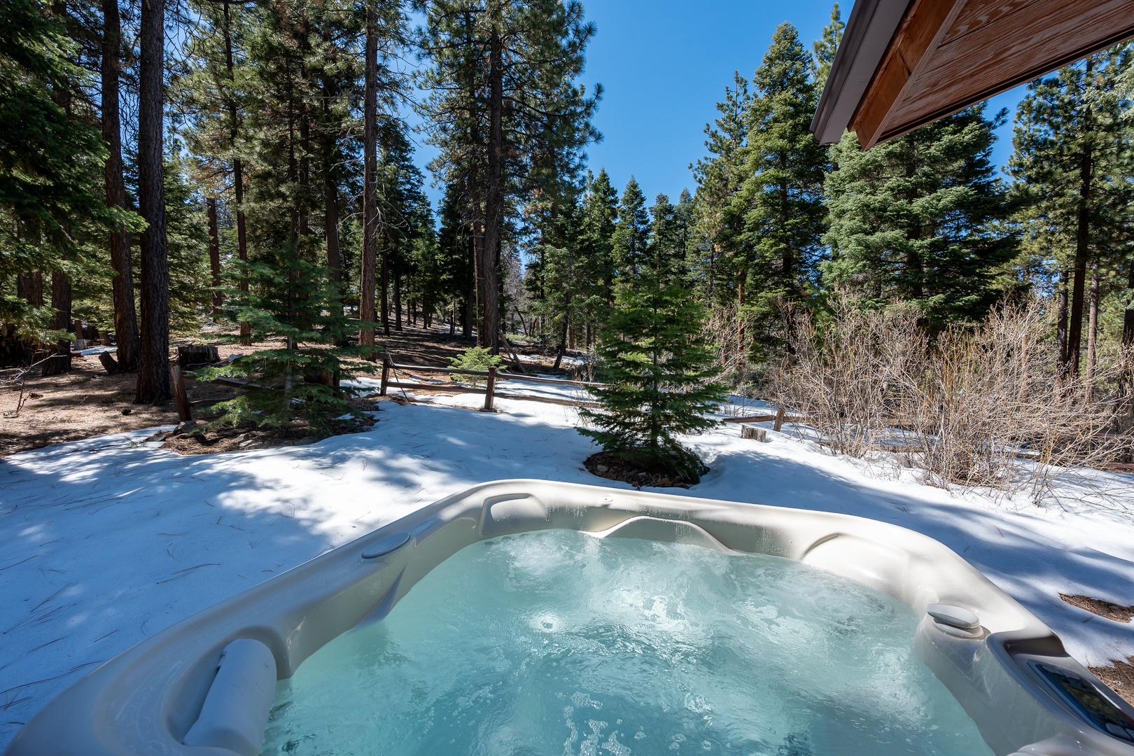 A steaming hot tub sits in a pristine winter setting surrounded by towering pines, creating a perfect alpine retreat where you can soak under clear skies.