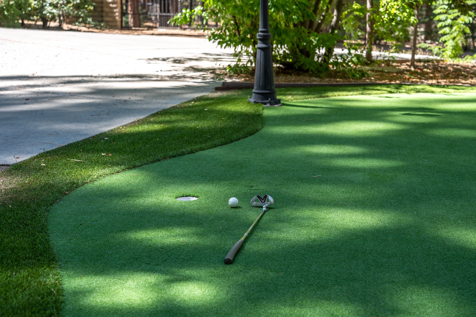 A miniature golf course set on smooth artificial turf, complete with a putter and ball ready for your next shot. The surrounding shade and natural greenery create a relaxing outdoor play area.