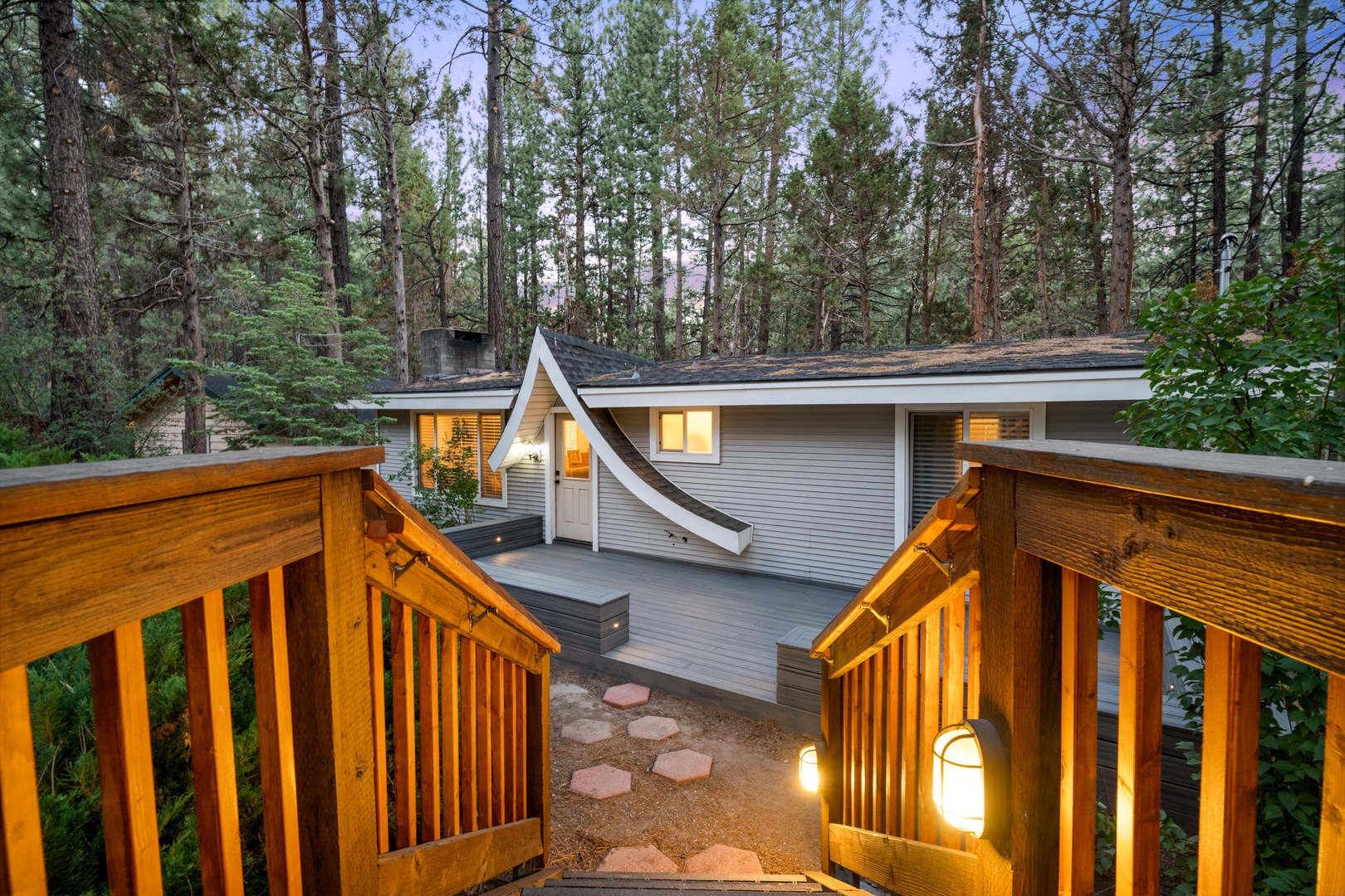 Nestled among towering pines, the property sits on peaceful wooded grounds. A wooden stairway with warm lighting leads to the cottage's entrance and private deck areas.