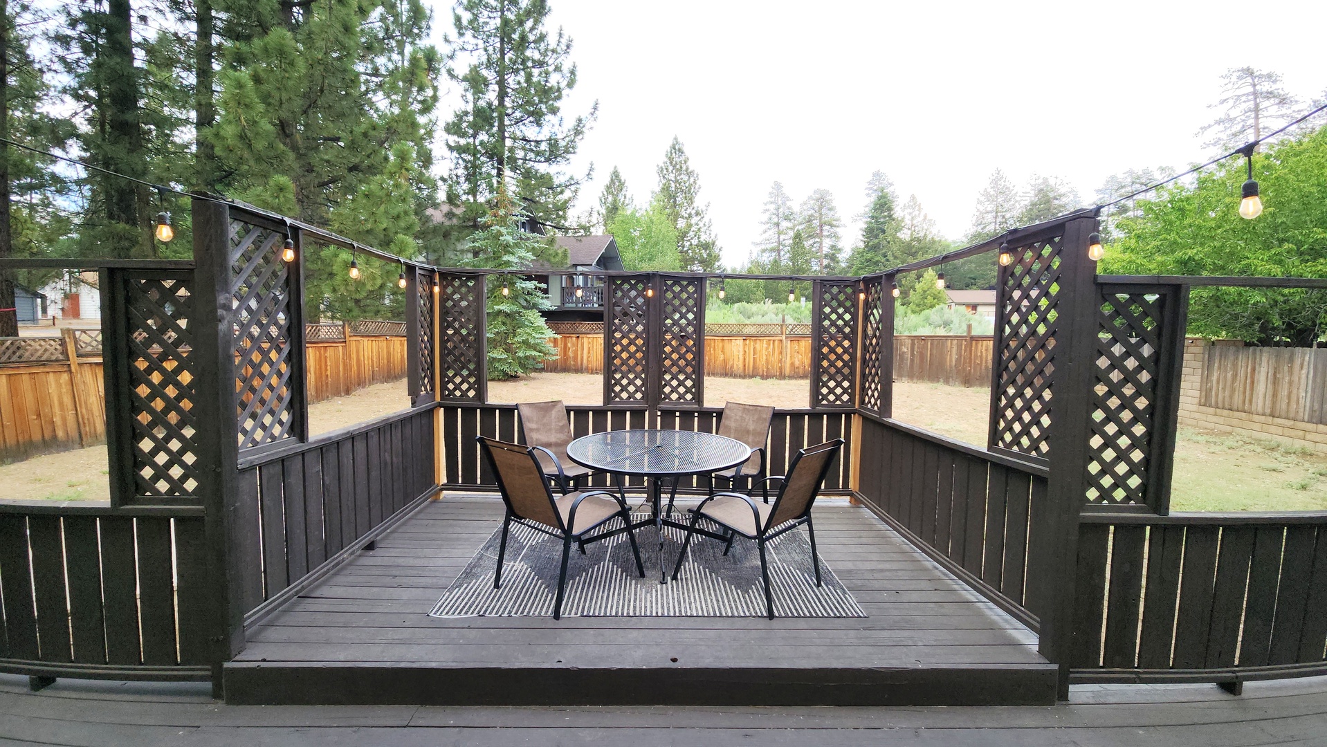 Your private outdoor dining deck features latticed privacy screens, string lights, and a cozy table set for four, perfect for meals surrounded by towering pines.