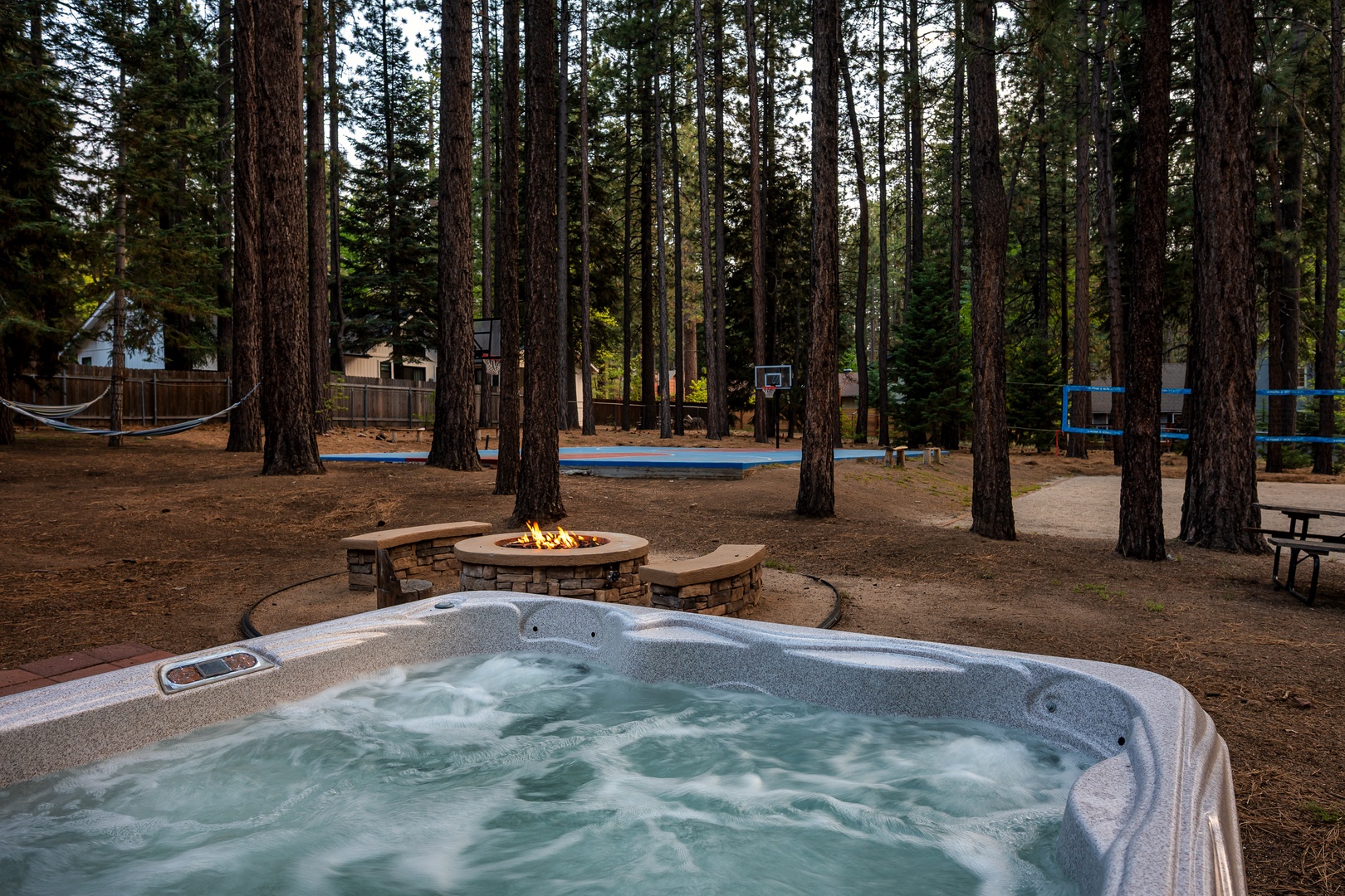 The property is nestled among towering pines, featuring a private hot tub, stone fire pit with seating, and recreational amenities including a basketball court and trampoline.