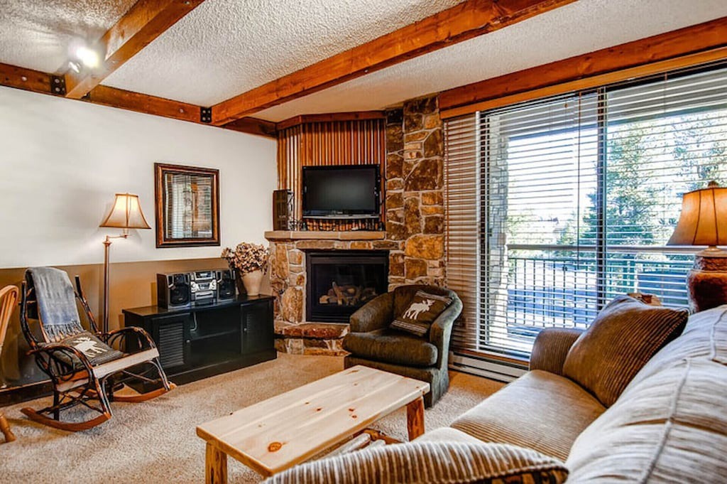 Cozy up by the stone fireplace in this warm living room featuring exposed beams and mountain views through expansive windows.