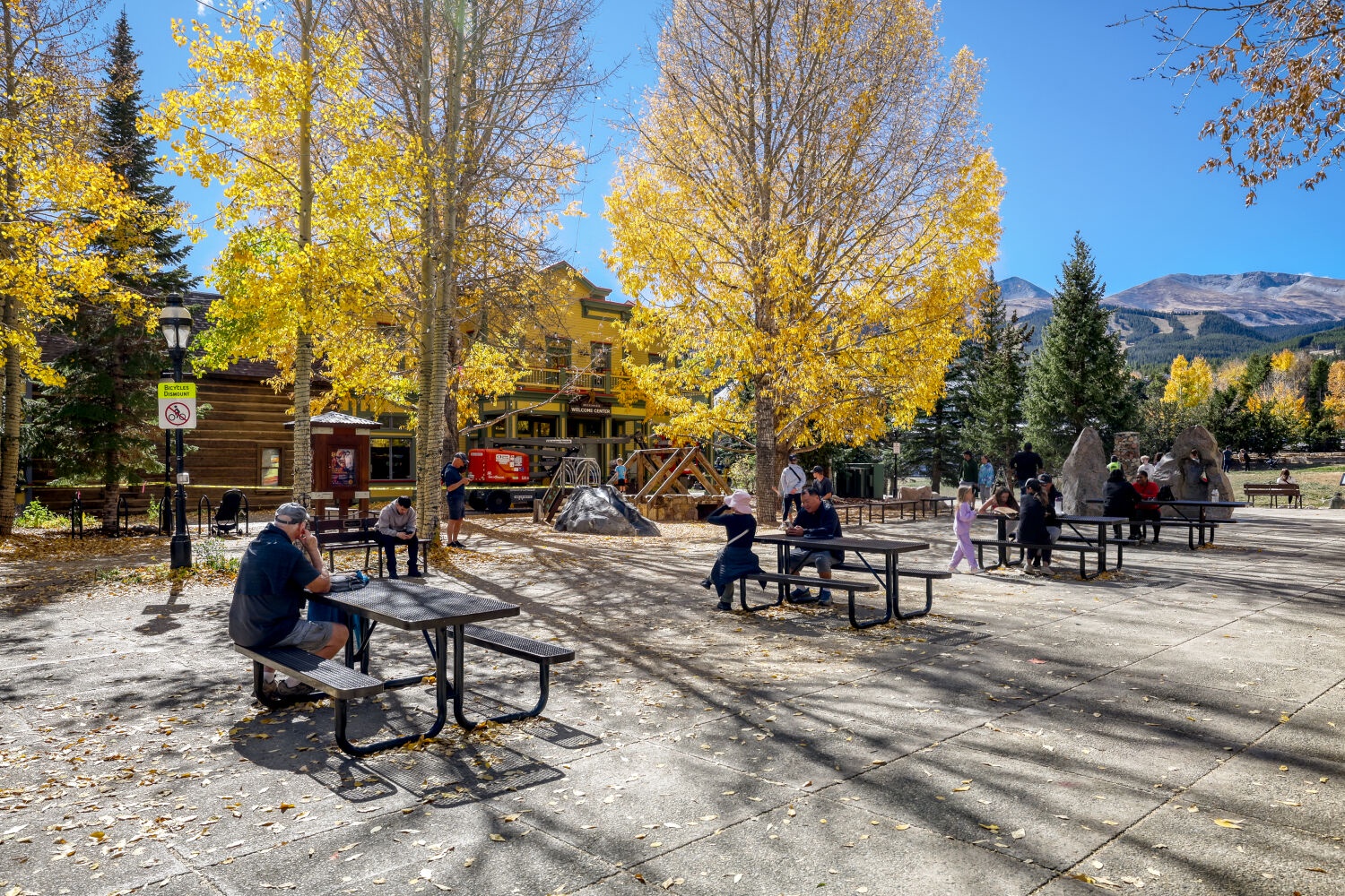 Breckenridge's Main Street area features picnic tables beneath golden aspens with distant mountain peaks framing the historic downtown backdrop. Captured on a sunny autumn day near local restaurants and gathering spots.
