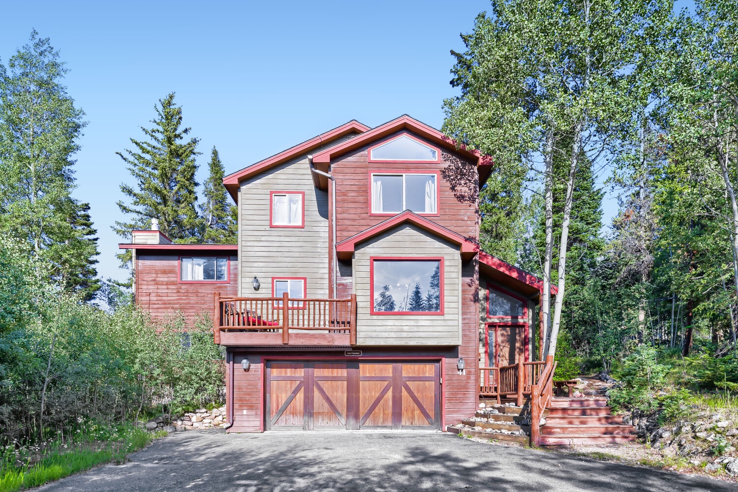 A charming mountain retreat nestled among towering evergreens, featuring warm red and gray tones, multiple balconies, and a welcoming entrance framed by natural stone steps and lush forest surroundings.