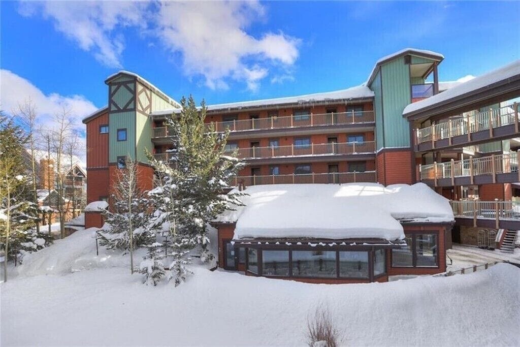 Snow-covered mountain lodge features distinctive architecture with green and red siding amid pristine winter landscape.