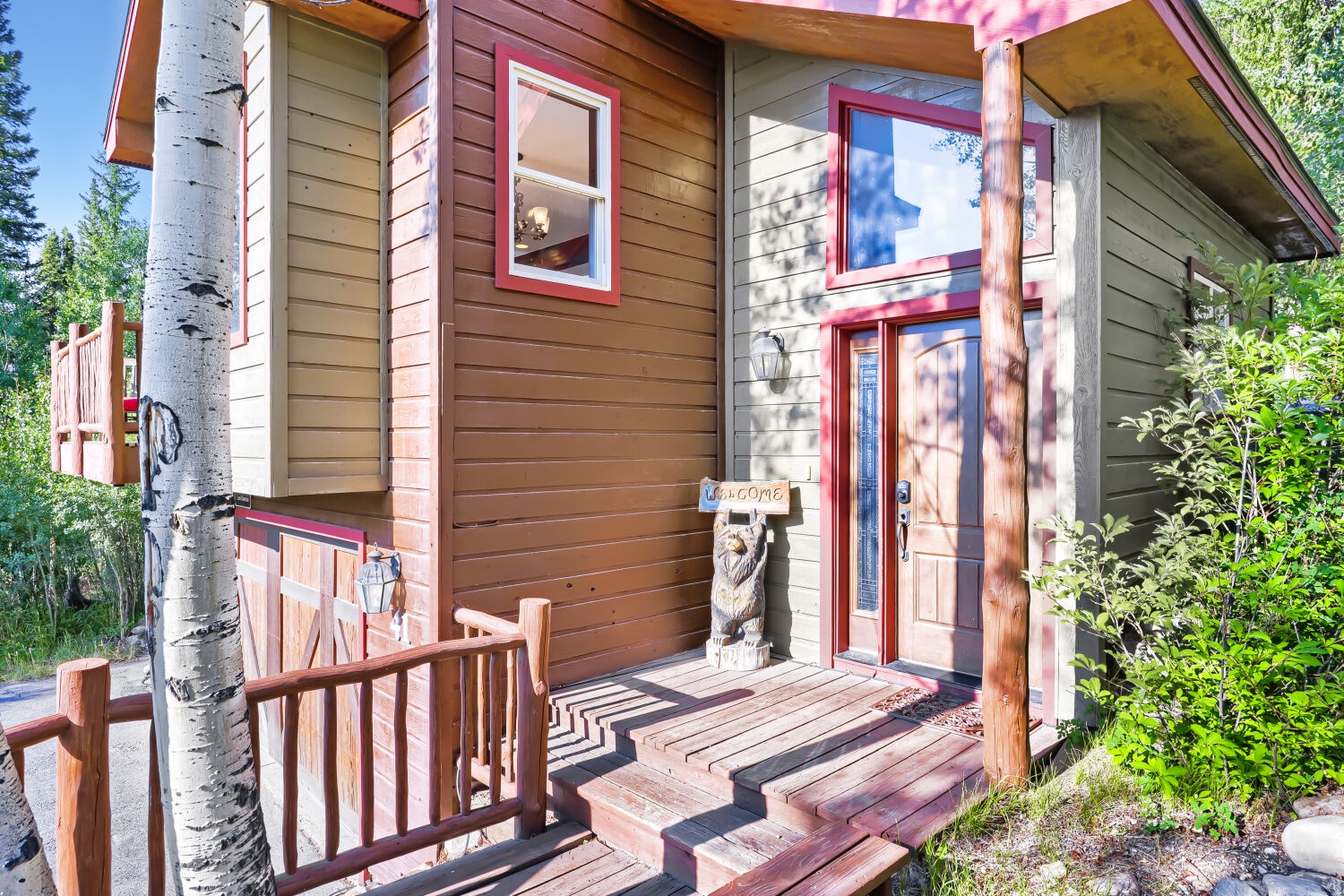 Charming mountain cabin entrance surrounded by aspen trees and natural greenery. The wood-carved bear greets visitors at the welcoming front door with its rustic wooden deck and stairs.