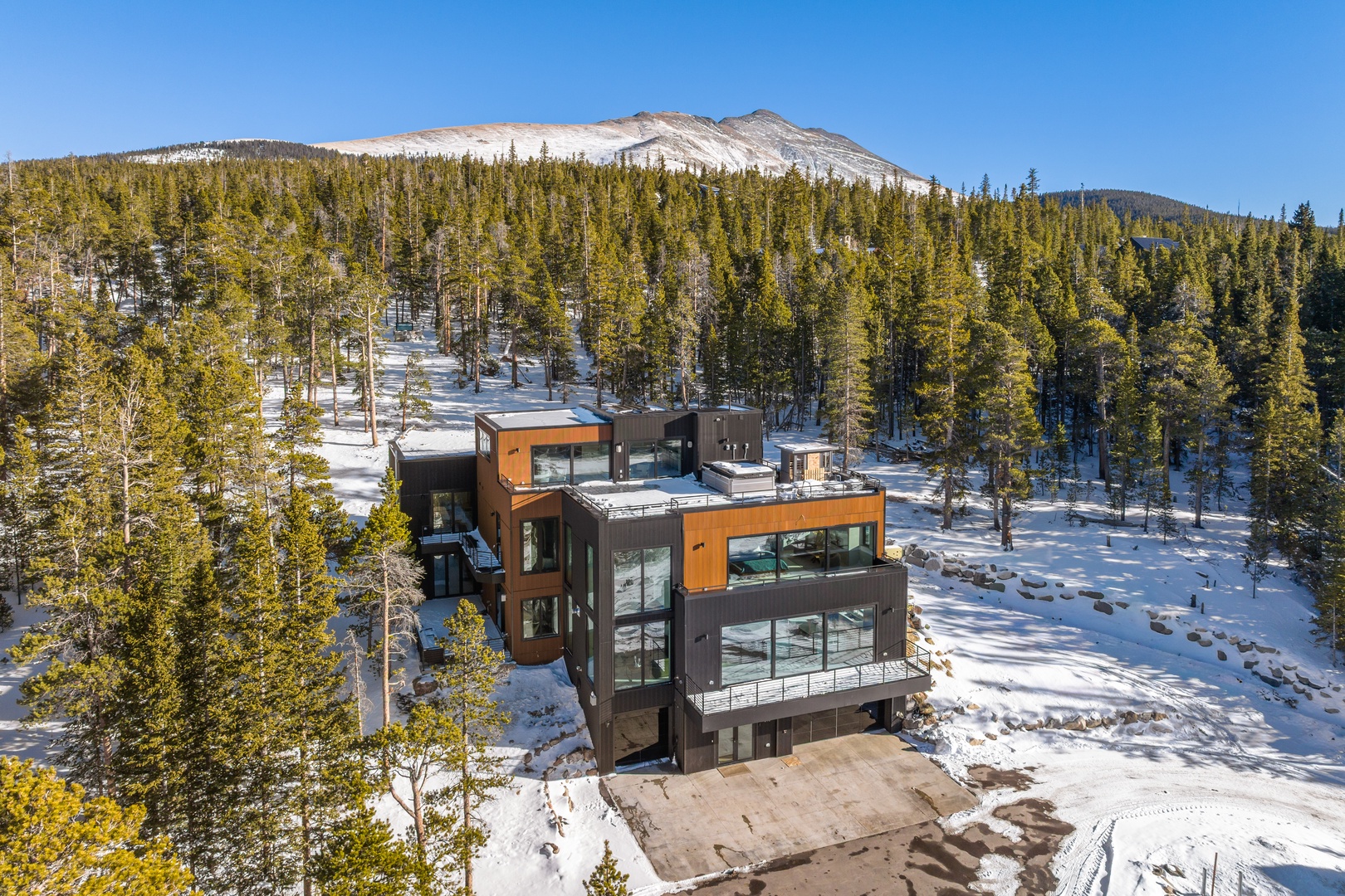 Modern mountain retreat nestled among snow-covered pines with dramatic alpine peaks rising in the distance.