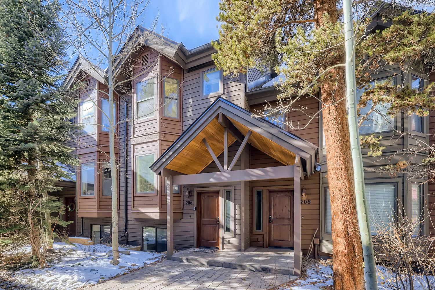 Mountain-style duplex with modern wood siding and covered entrance, surrounded by mature evergreens and winter landscape.