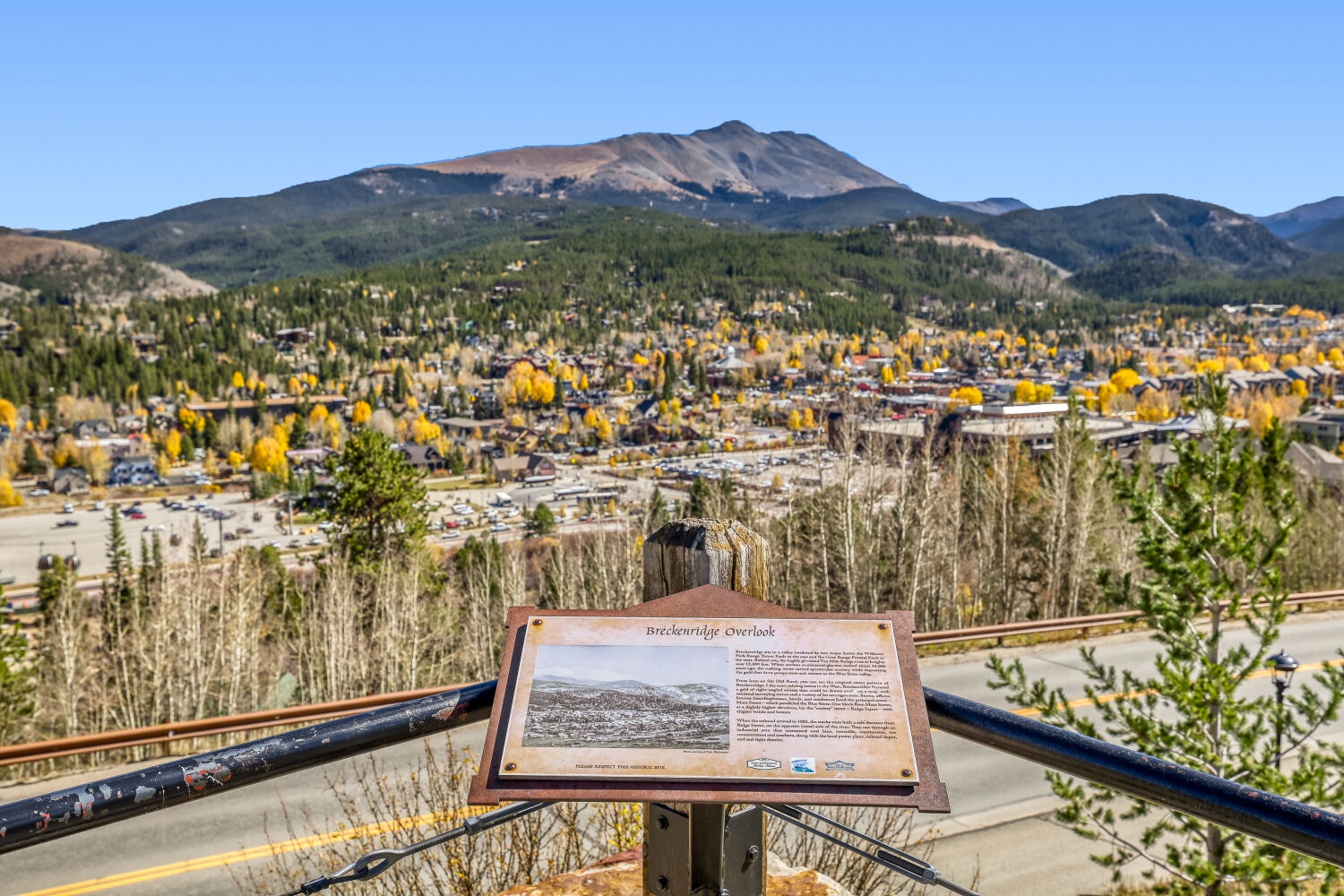 A scenic overlook offers expansive views of the mountain town nestled below, framed by golden autumn foliage and dramatic peaks stretching across the horizon.