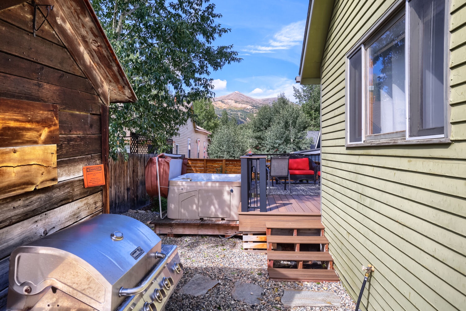 This charming mountain retreat features a private hot tub, gas grill, and deck nestled between rustic buildings, with stunning mountain peaks framing your outdoor sanctuary.