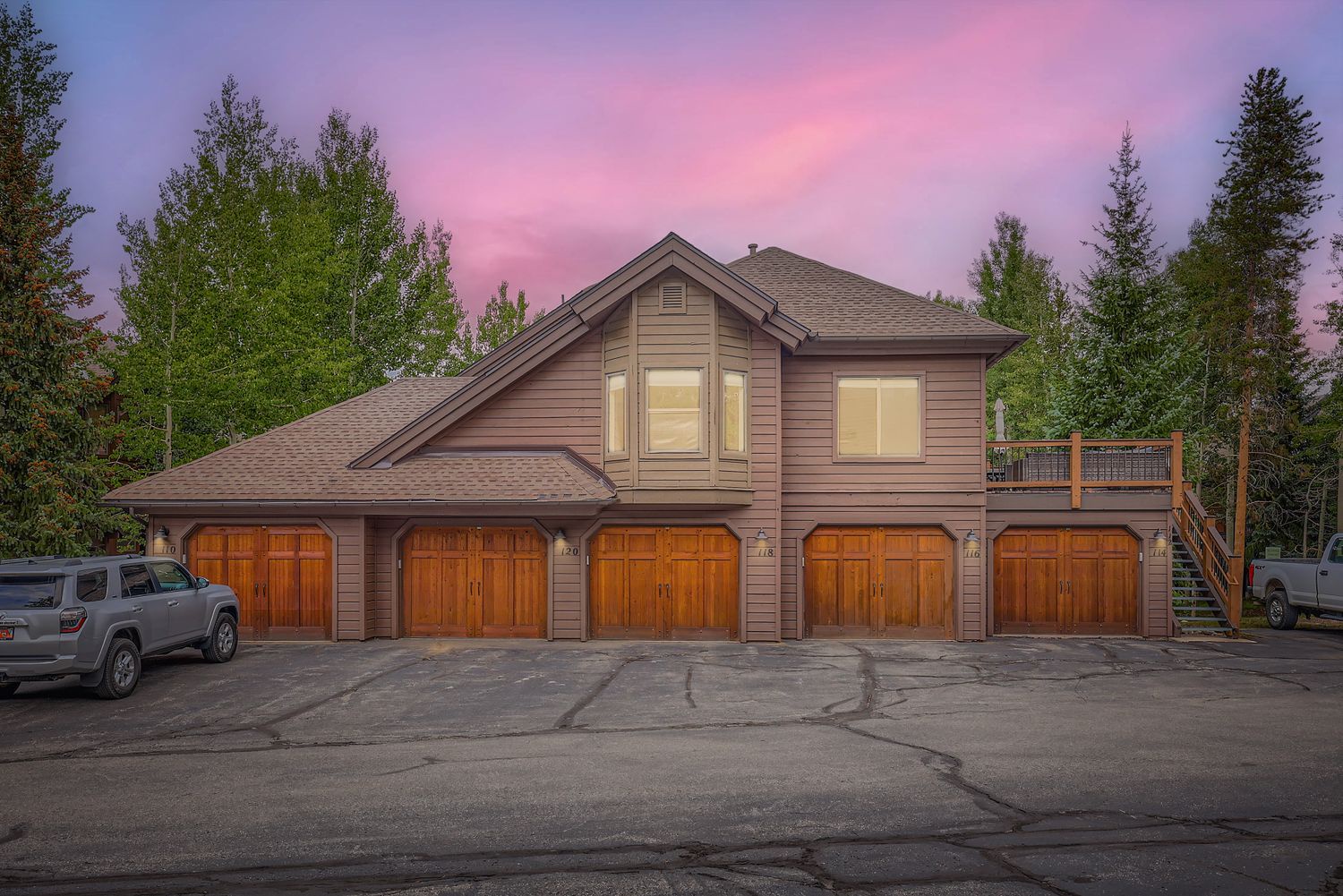 This charming mountain lodge building features warm wood siding and multiple garage spaces, nestled among towering evergreen trees under a stunning twilight sky.