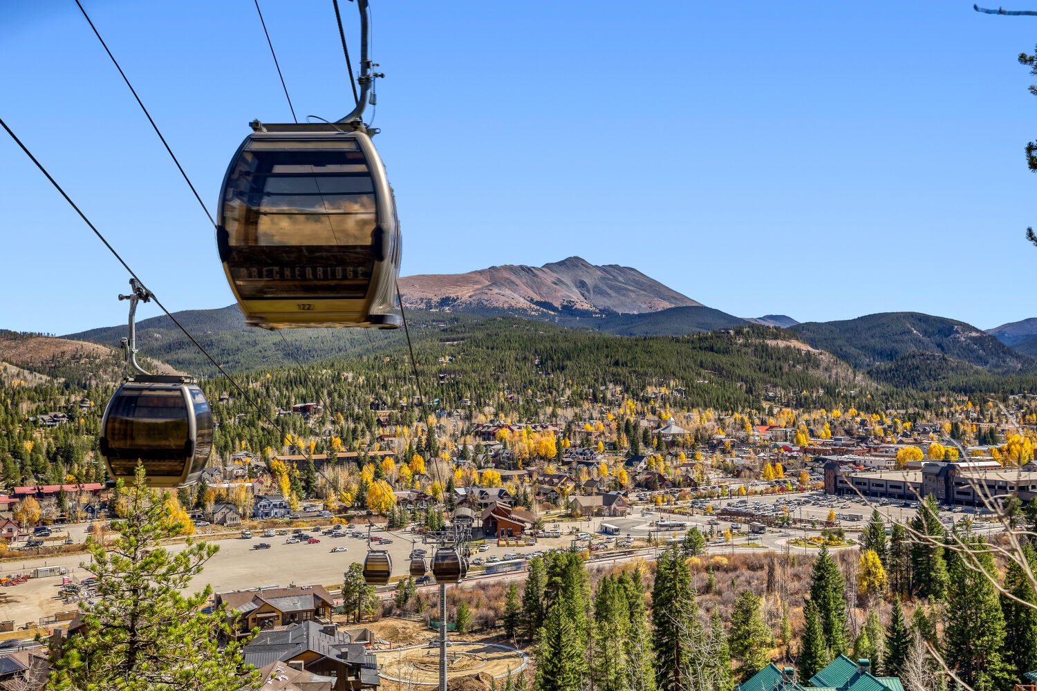 A scenic gondola glides above a mountain town nestled among evergreens and golden aspens, with dramatic peaks rising in the background under clear autumn skies.