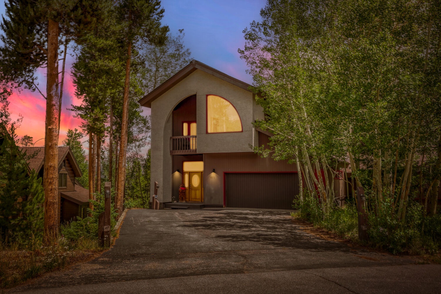 A striking modern home nestled among tall pines, illuminated at twilight with a spectacular sunset painting the sky in soft pink and orange hues—an inviting arrival to a tranquil mountain escape.