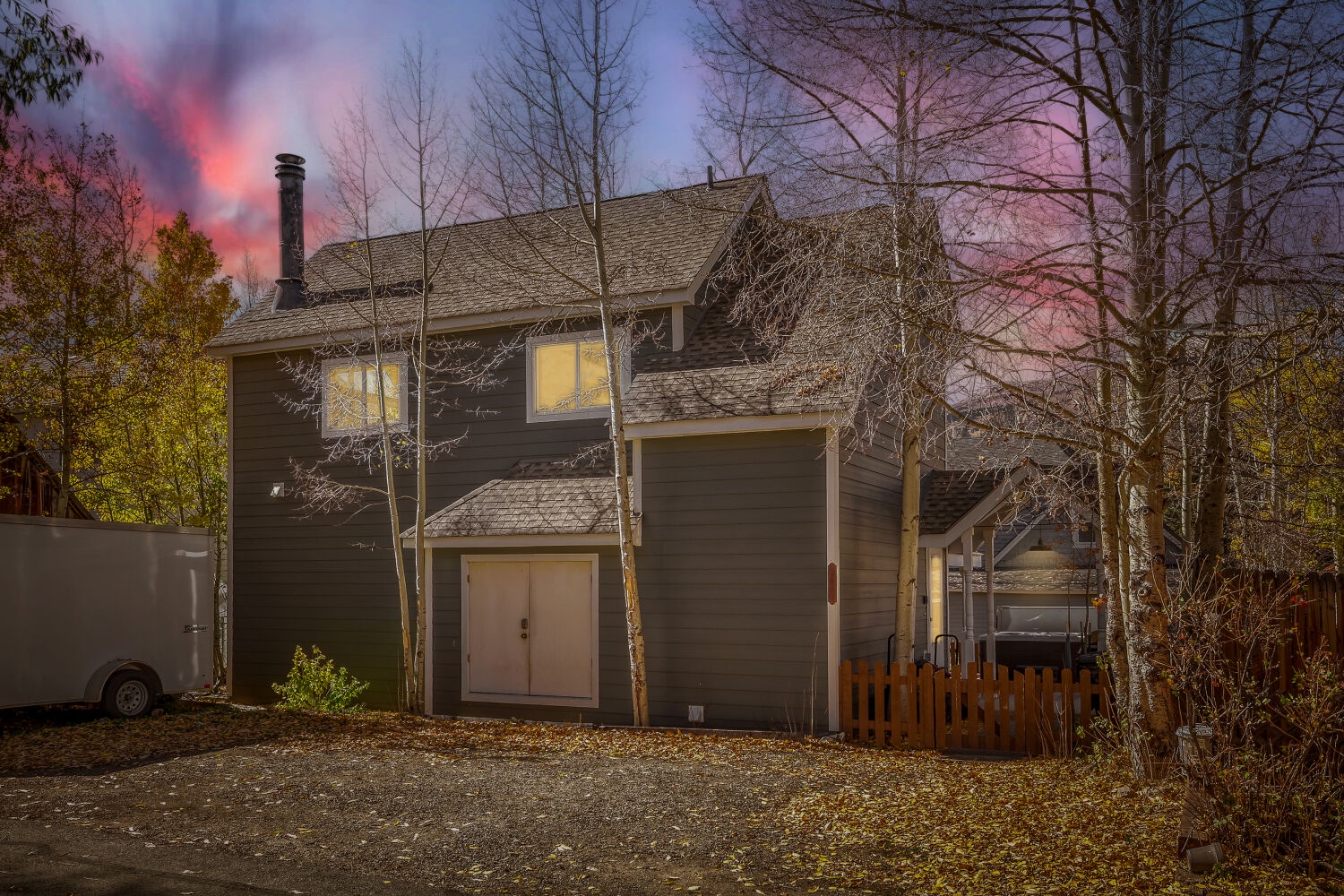 A charming mountain retreat framed by aspens and evergreens, captured at dusk beneath a dramatic pink and lavender sky, with warm light glowing from the windows.