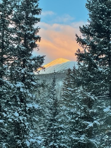 Snow-dusted evergreens frame a stunning alpenglow sunrise over nearby mountain peaks, captured from the property's scenic surroundings.