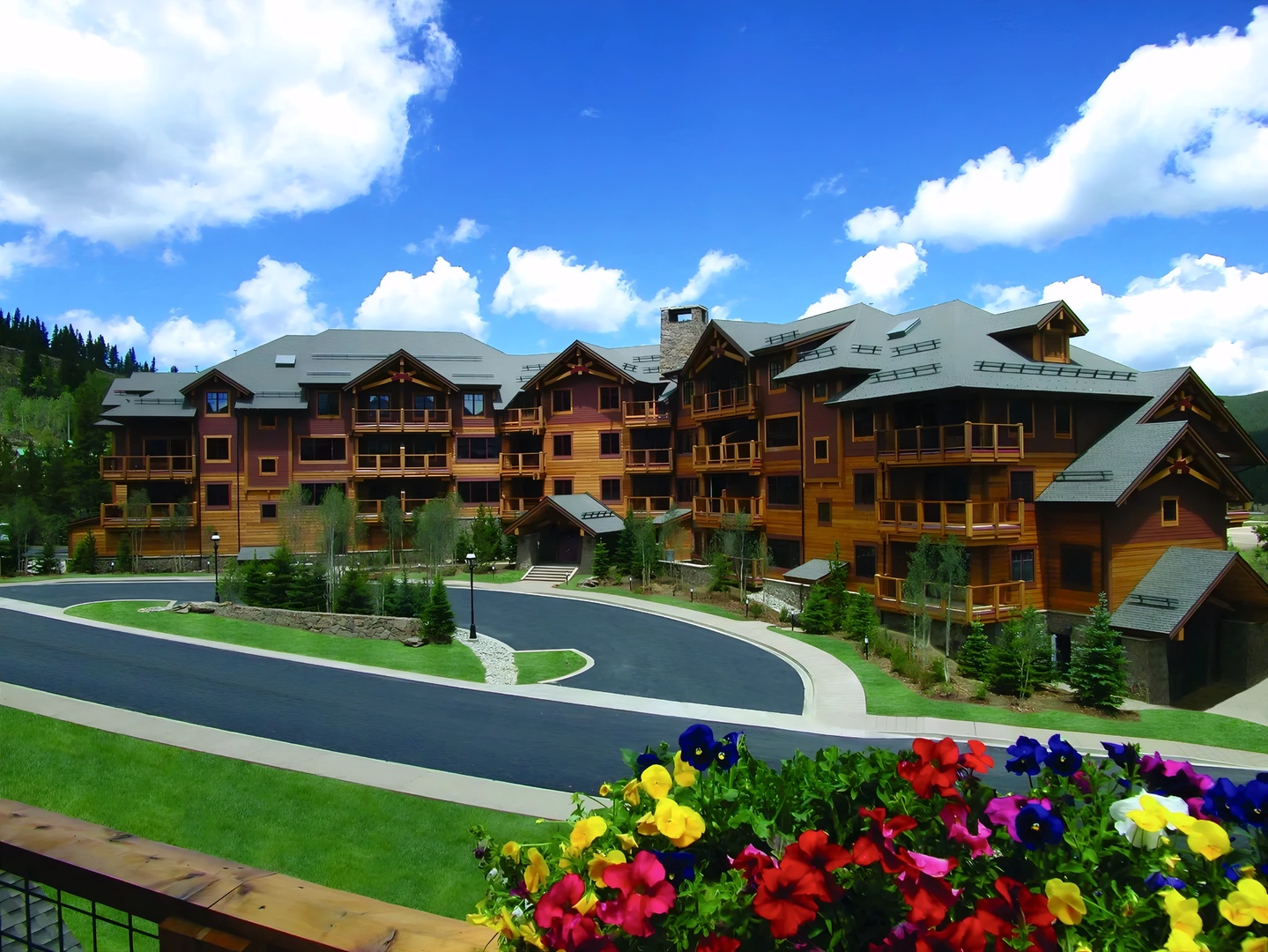 Mountain lodge complex featuring rustic alpine architecture with colorful flower beds and curved entrance roadway.