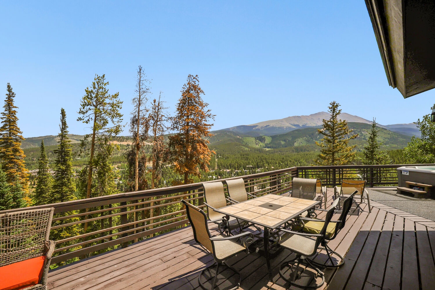 Spacious wooden deck with outdoor dining beneath expansive mountain and forest vistas, bathed in blue-sky sunshine.