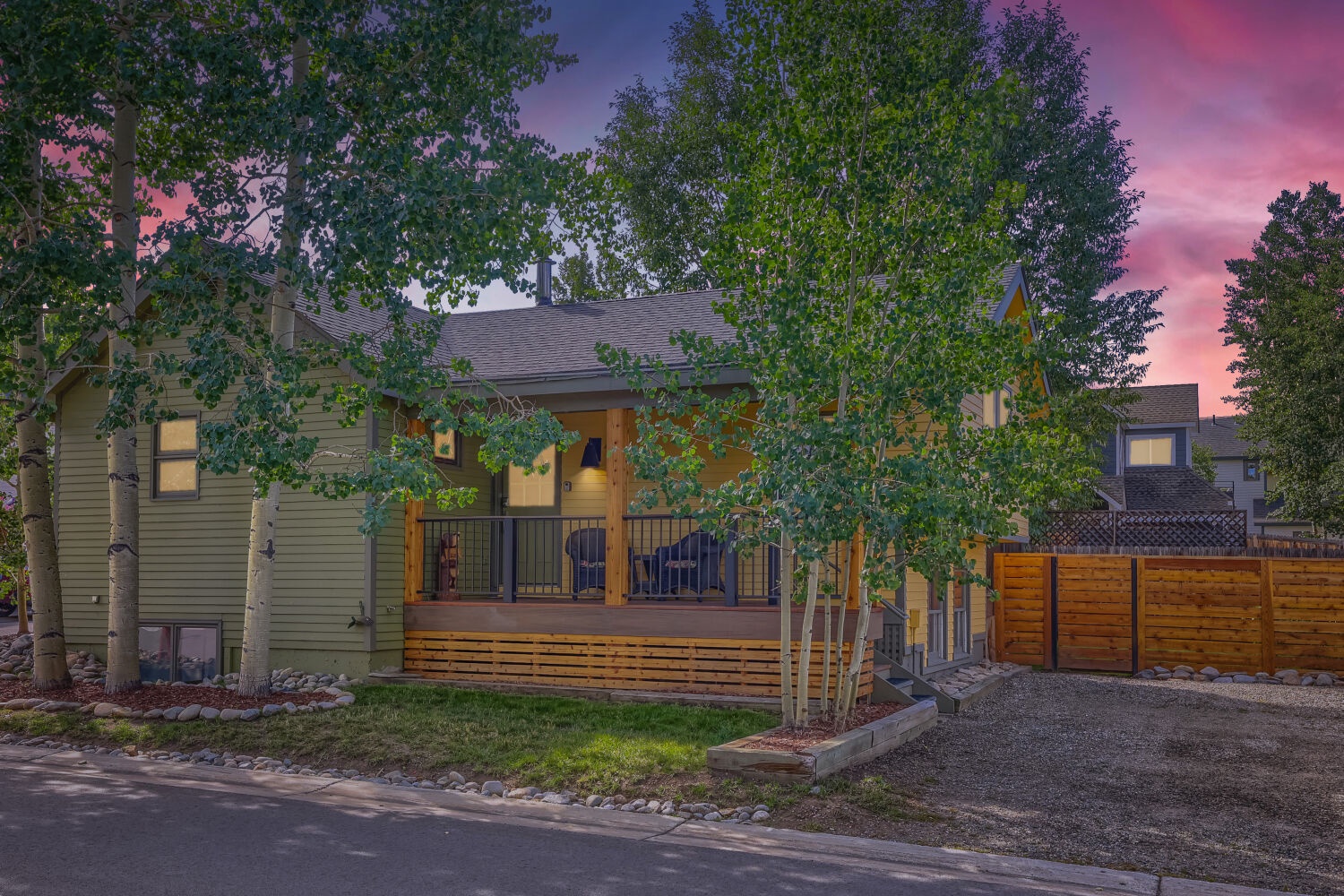A charming property nestled among mature trees, featuring a welcoming covered porch and warm outdoor lighting against a stunning pink and purple twilight sky.