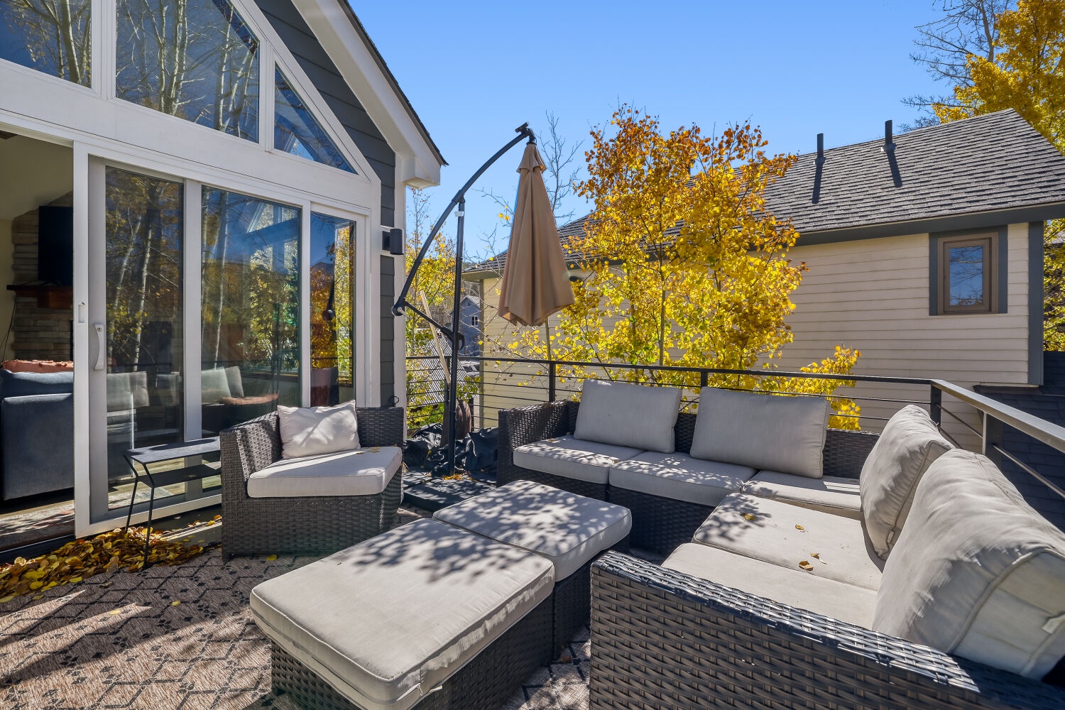 This spacious sun deck features comfortable wicker seating with plush cushions, perfect for enjoying crisp autumn days with a warm drink. The dappled shade and vibrant foliage create a peaceful retreat for your relaxation.