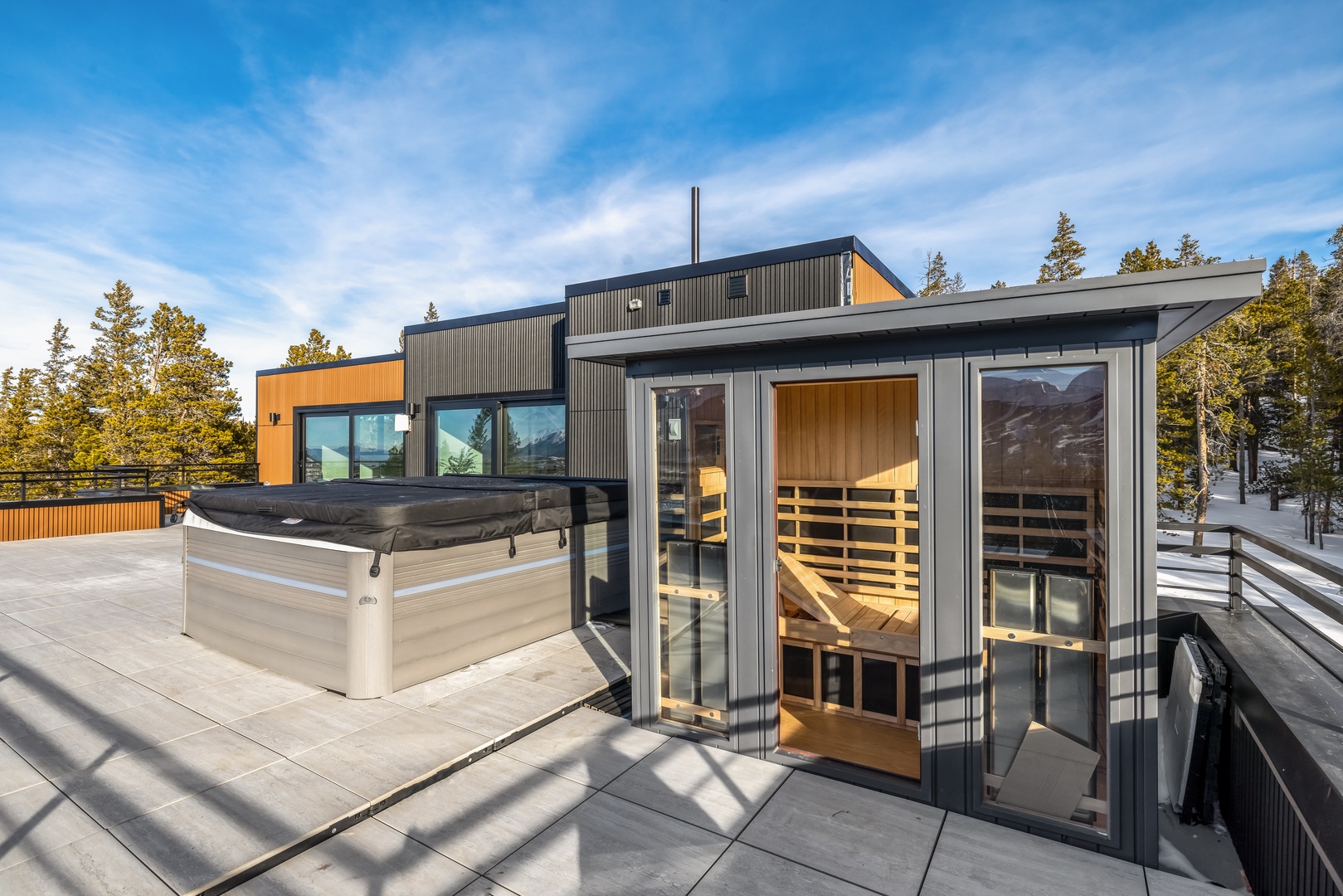 Modern mountain retreat features outdoor sauna and hot tub on spacious deck surrounded by snow-dusted pines.