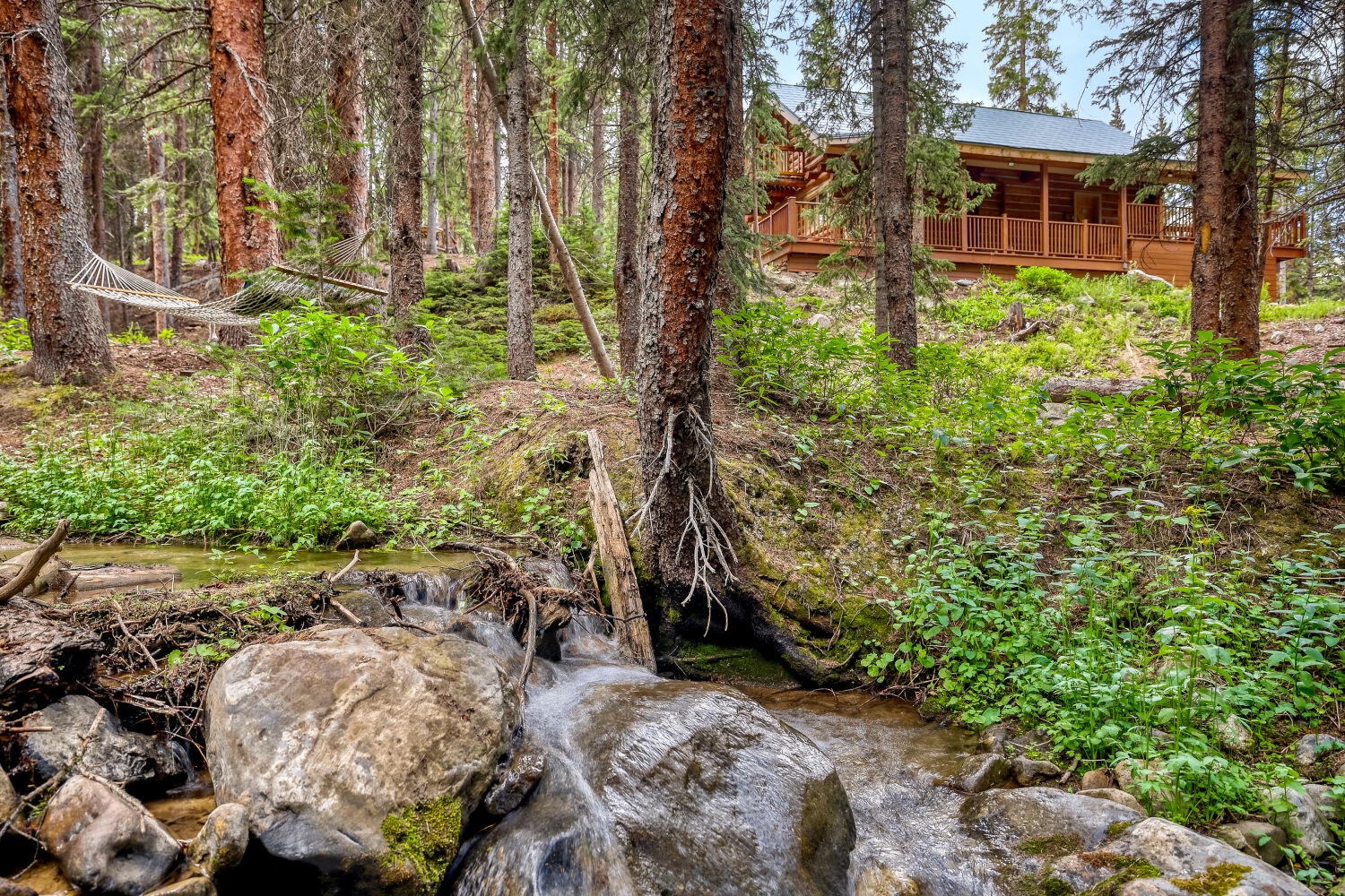 A warm log cabin nestled among towering pines alongside a gently flowing forest stream, providing a peaceful natural backdrop for your mountain getaway.
