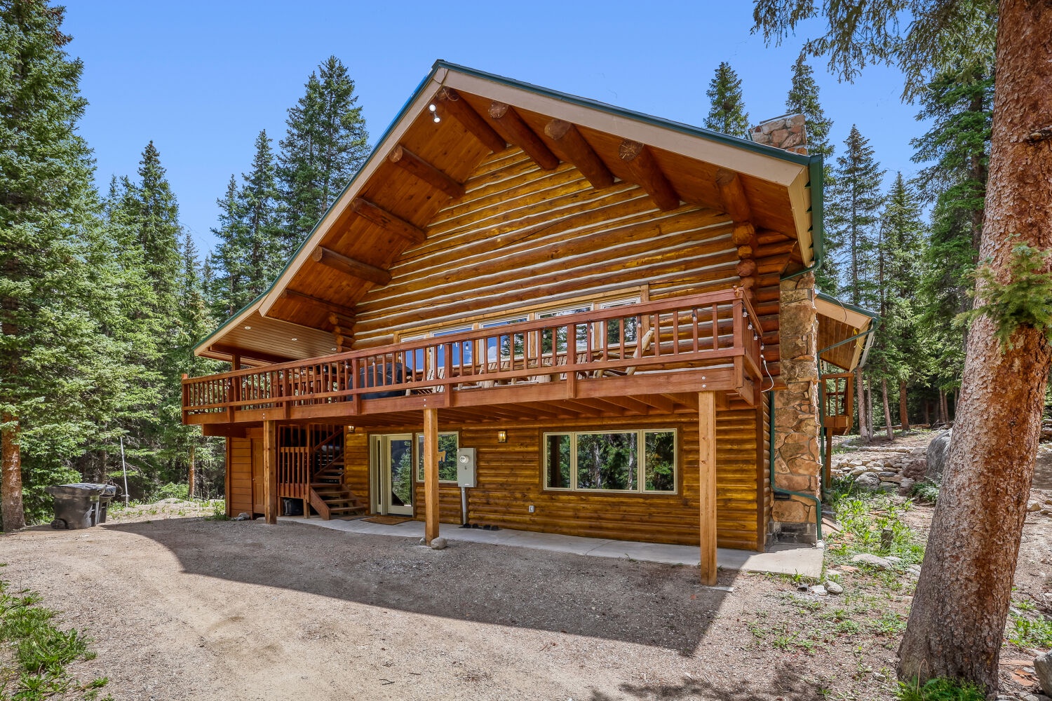 A charming log cabin nestled among towering pines, featuring a welcoming balcony and rustic wood exterior. The property sits within a peaceful forest setting with ample parking and outdoor space.